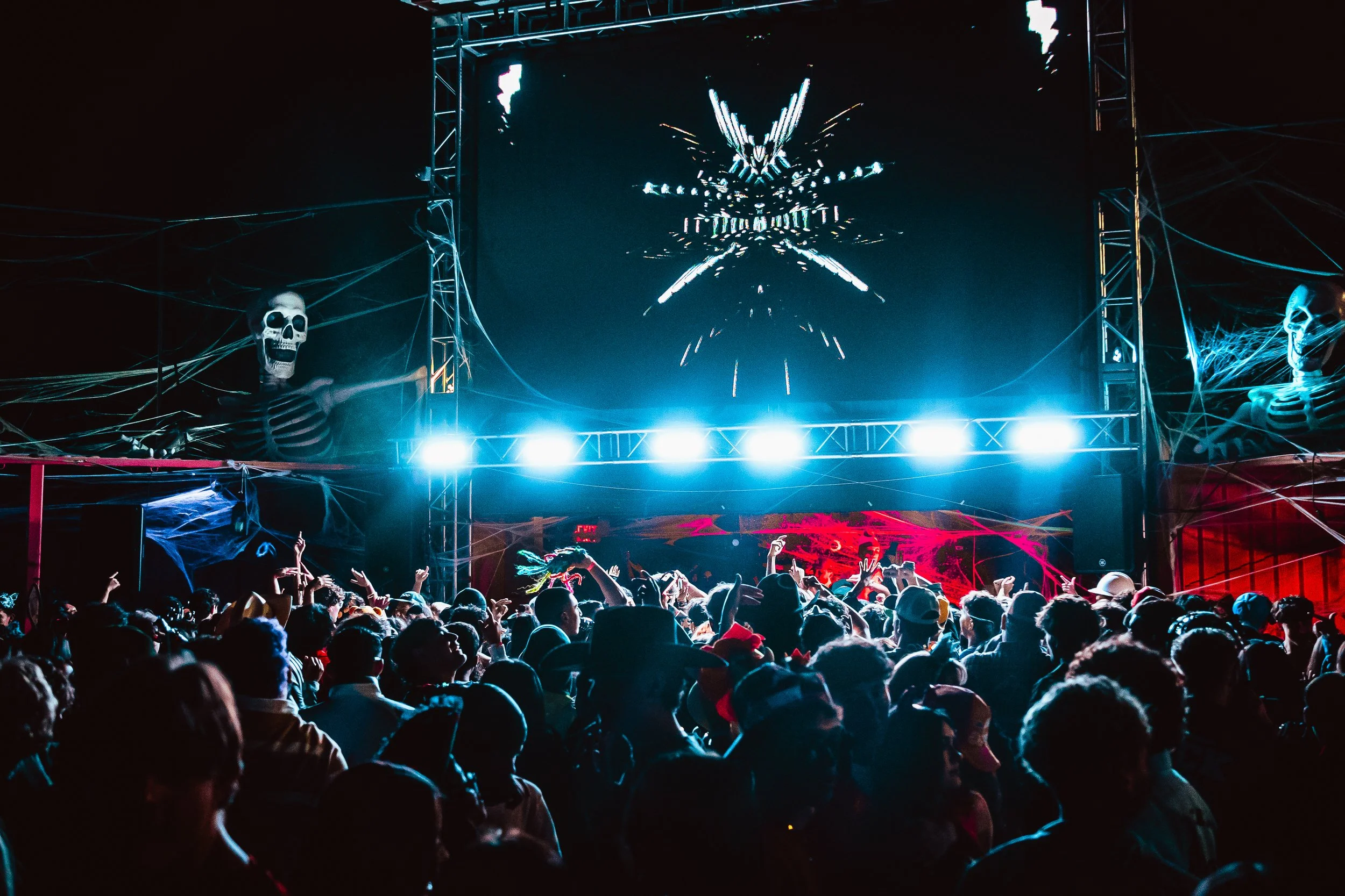 Crowd at a Halloween-themed concert or event with skeleton decorations, web-like embellishments, and a large screen displaying abstract visuals.