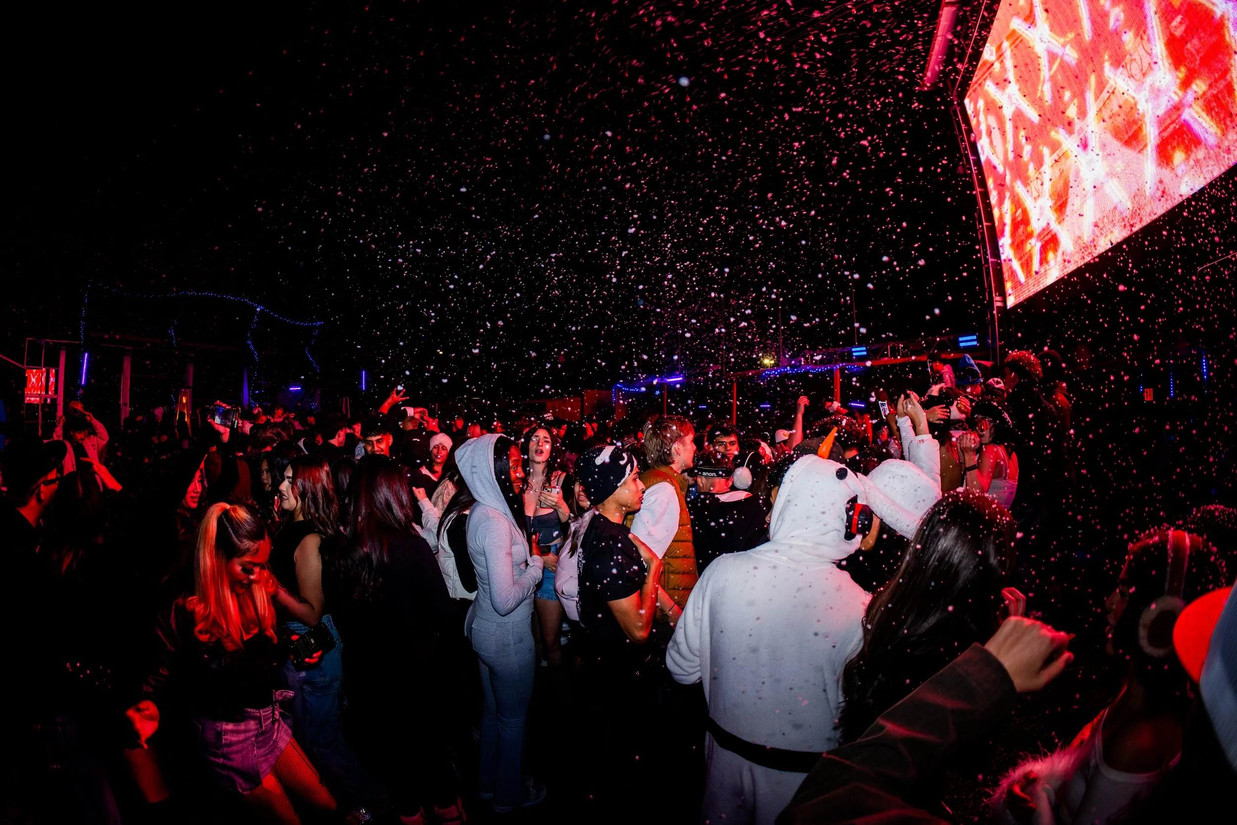 Crowd of people at a party or concert under red lighting, with snow-like confetti falling from the ceiling and a large screen displaying visuals.