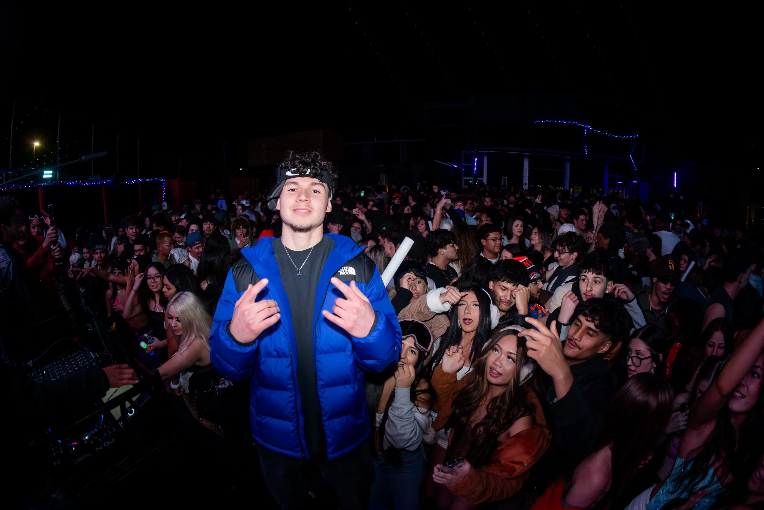 Young man in blue jacket and black headband standing in front of crowded dance party with people dancing and colorful lights