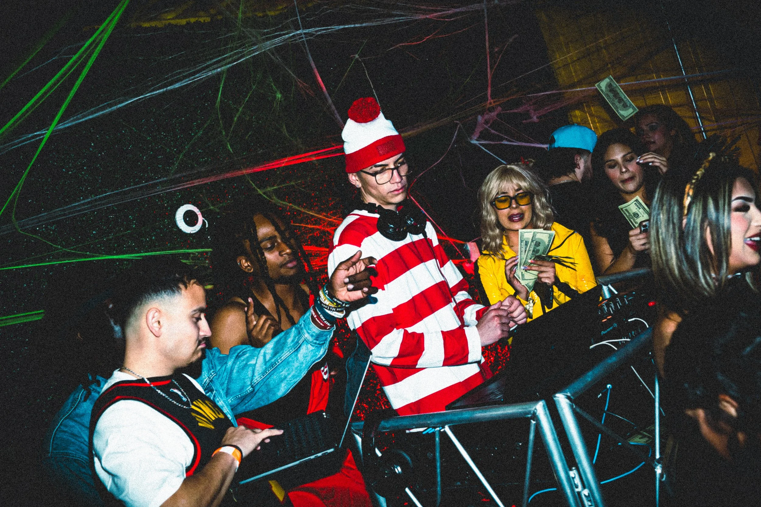 Group of young people dancing and enjoying a party at a nightclub with colorful laser lights, some holding cash, and one wearing a red and white striped shirt and a beanie.