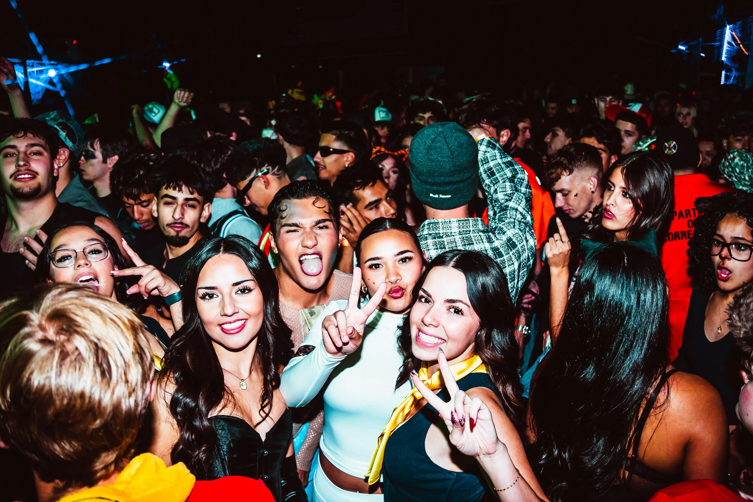 A large group of young people at a party or concert, some making peace signs, others posing for the camera, in a crowded indoor setting with colorful lighting.