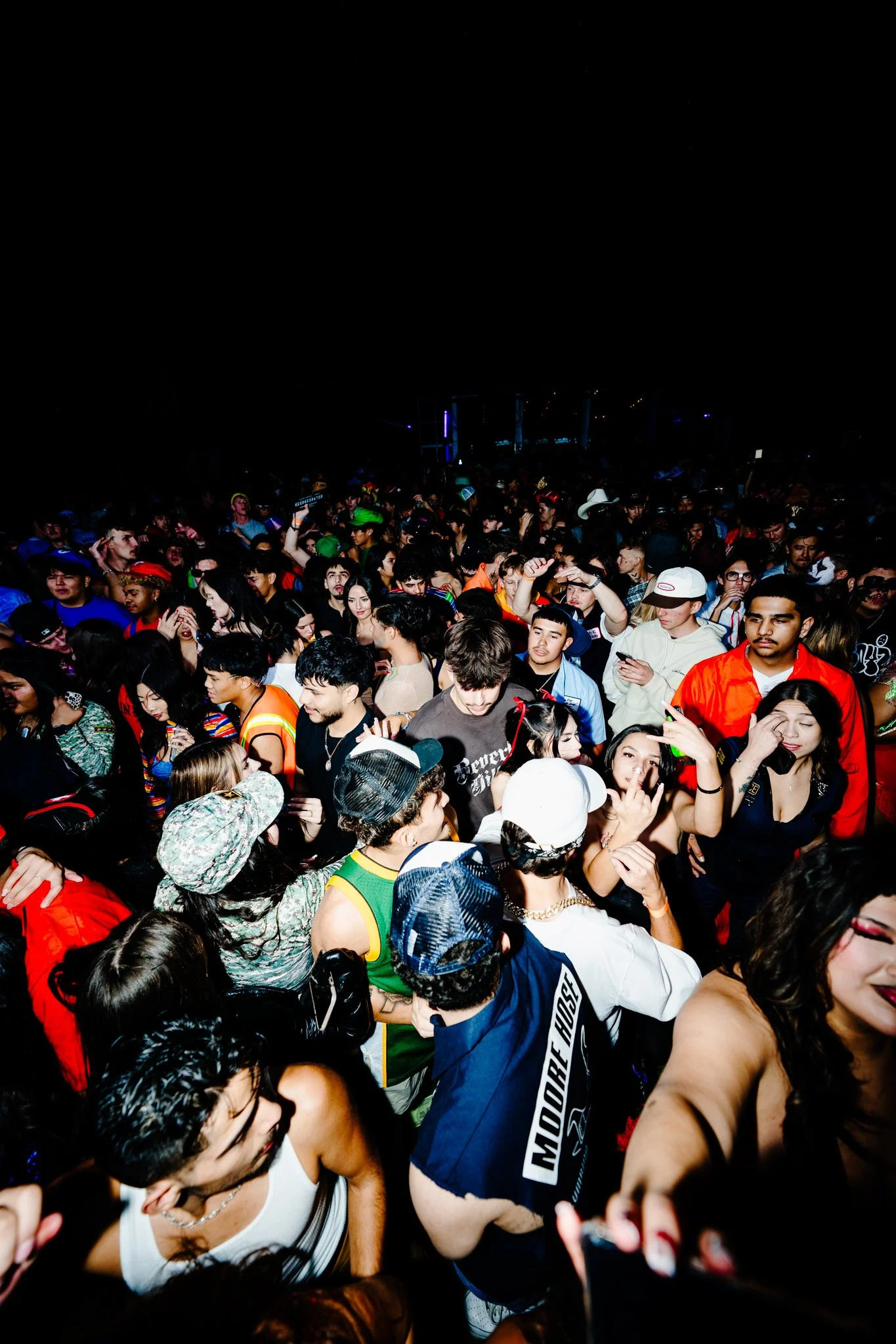 Crowd of diverse young people at a concert or nightclub dancing and socializing under dark lighting.