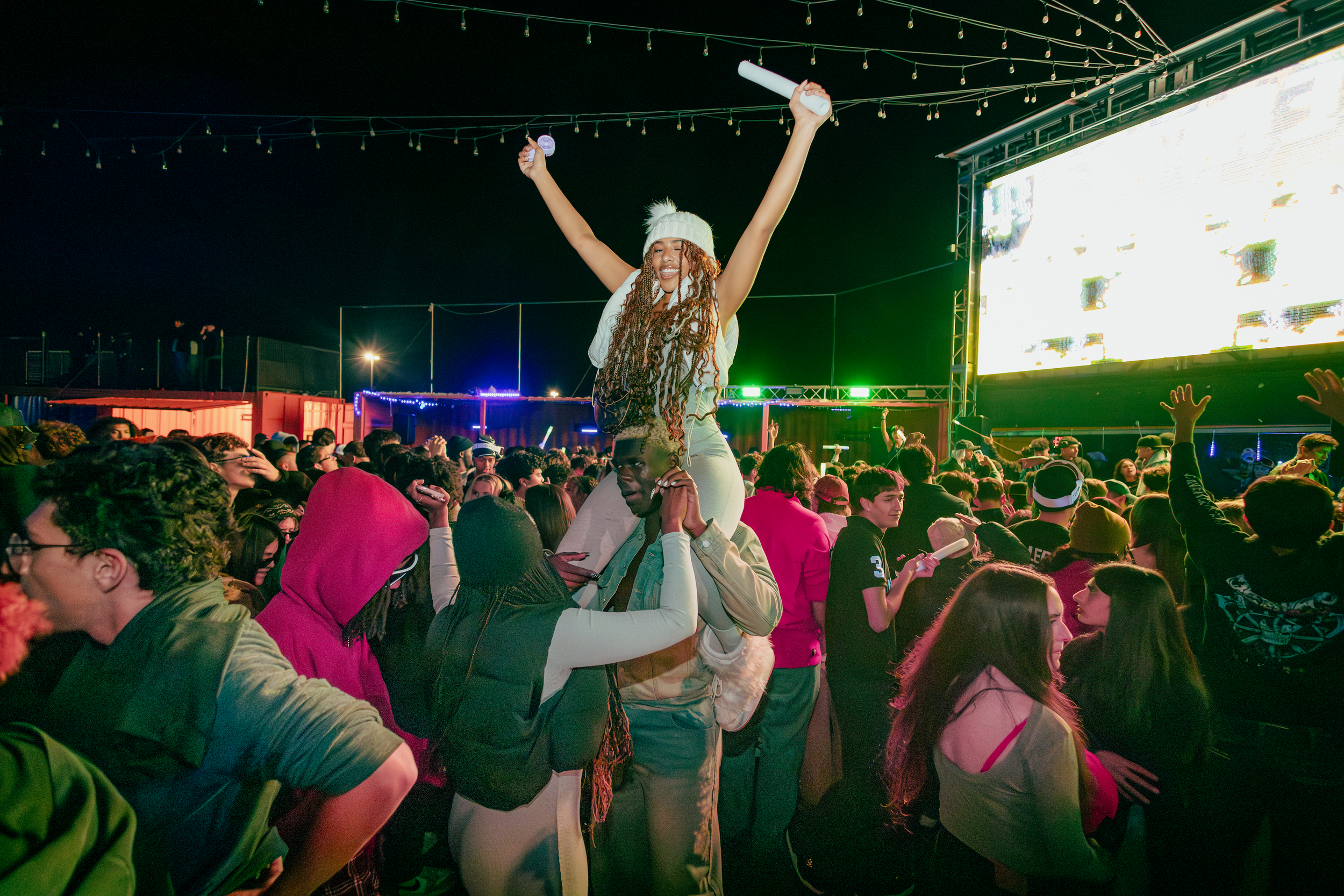 A large crowd at an outdoor concert or festival at night, with a performer being carried on someone's shoulders, raising arms in celebration, and a big screen to the right displaying visual effects.