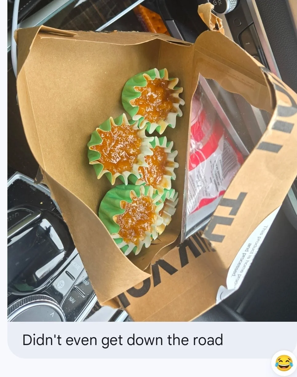 A paper bag with four cupcakes topped with caramel and green cupcake liners inside a car. The bag has black text and different items are in the car's interior.