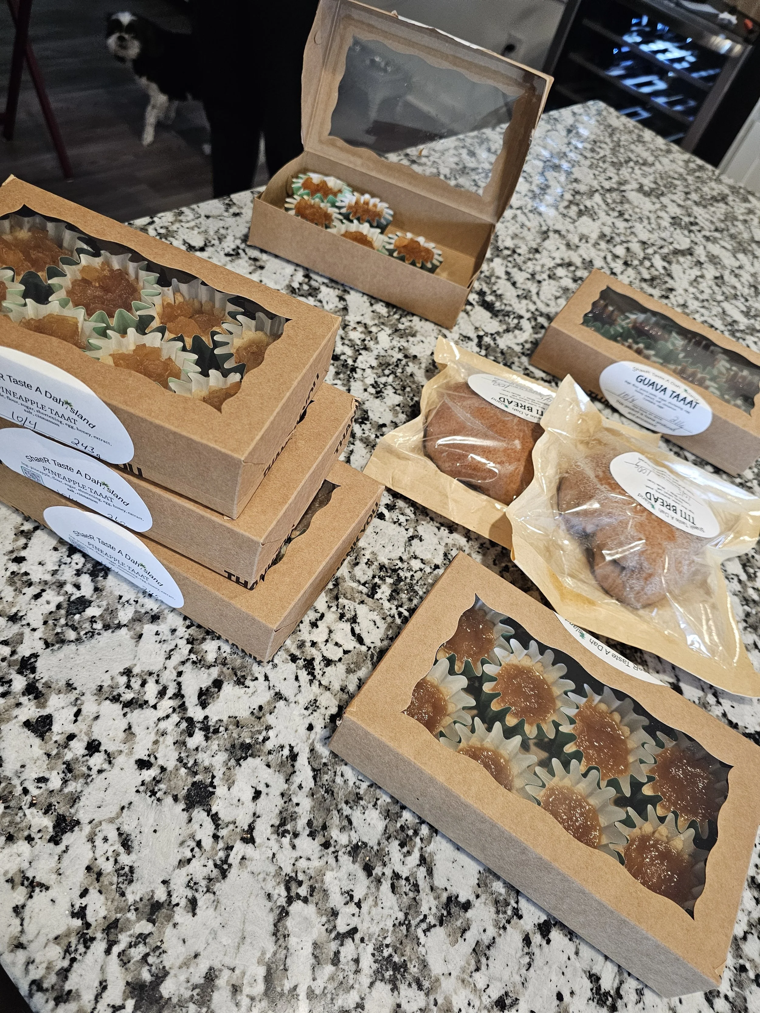 Box of homemade caramel candies, some in clear plastic bags and others in cardboard boxes with transparent windows, on a granite countertop in a kitchen with a dog in the background.