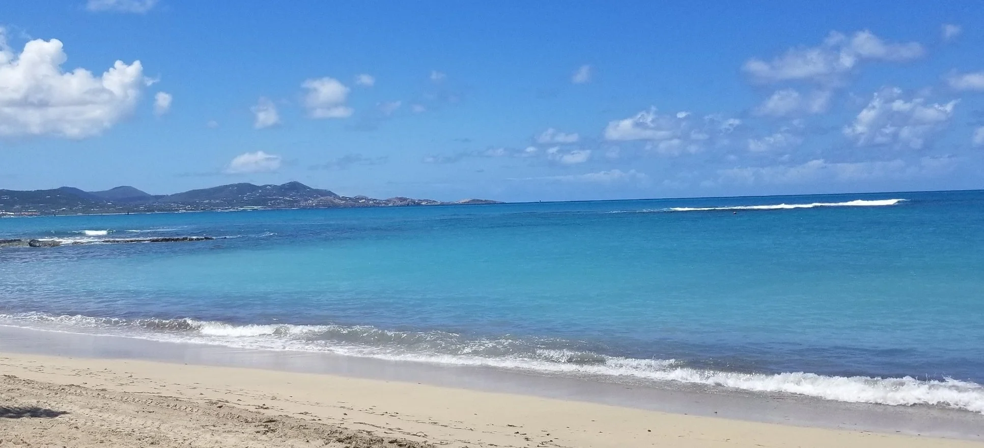 A tropical beach with white sand, clear blue water, and an expansive sky with fluffy white clouds. Distant mountains are visible on the horizon.