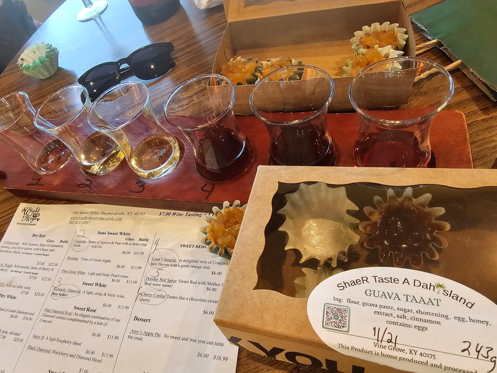 A wine tasting setup with six small glasses filled with different shades of wine placed on a wooden tray numbered 1 to 6. There are snack cups with some kind of pasty snack, a menu with wine descriptions, a pair of black sunglasses, a green cupcake l