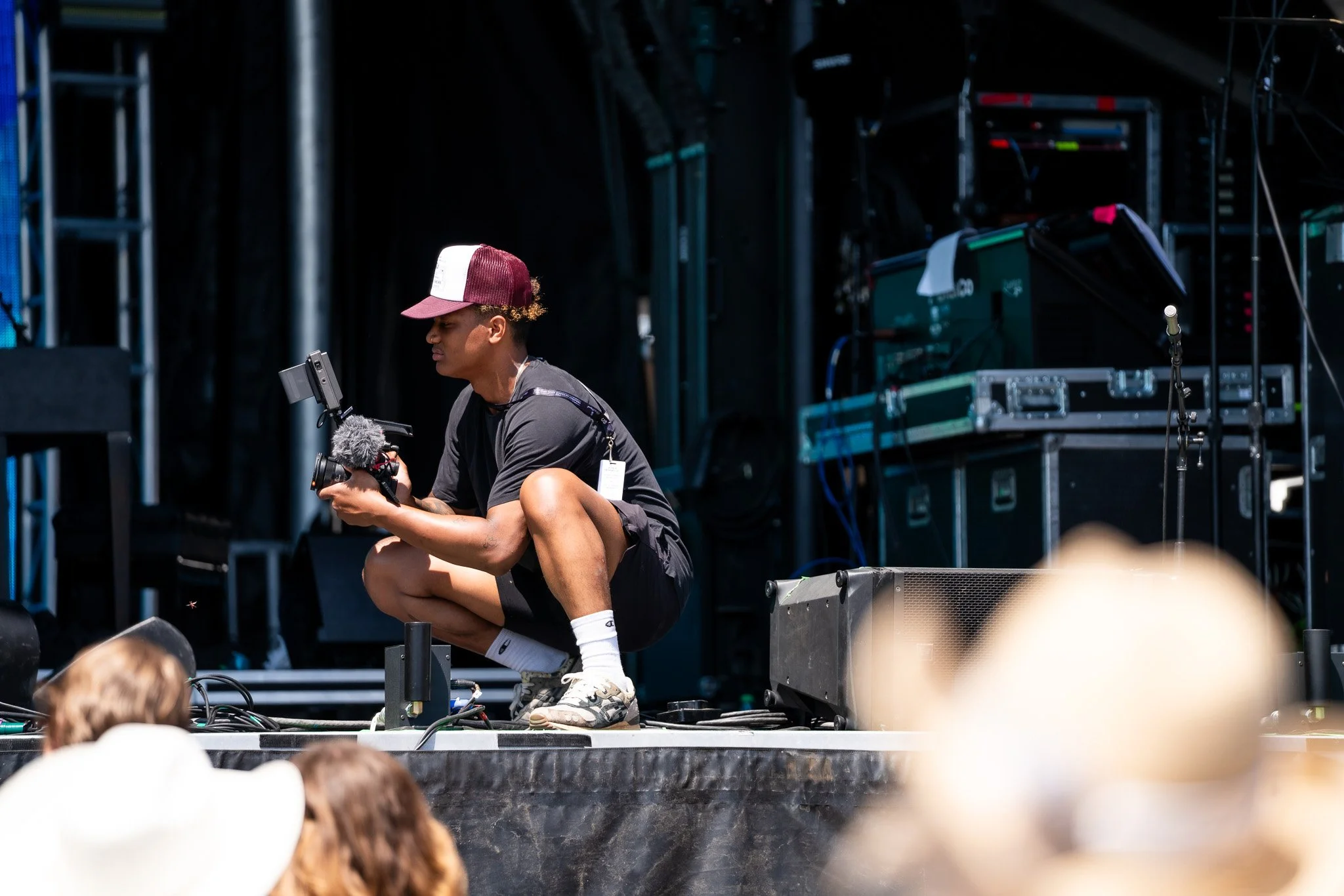 A young man squatting on a stage, holding a camera with a microphone, surrounded by audio and stage equipment, with audience members in the foreground.