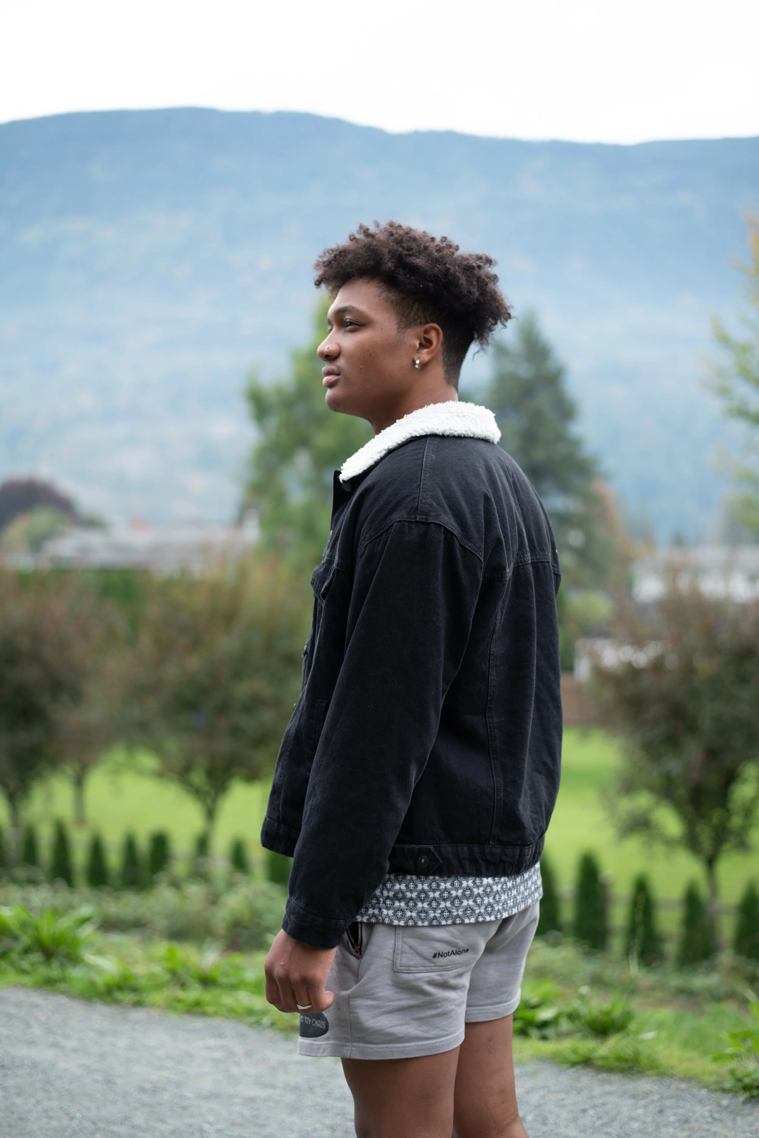 A young person with short curly hair standing outdoors in front of a mountain landscape, wearing a black jacket with a white collar and light-colored shorts.