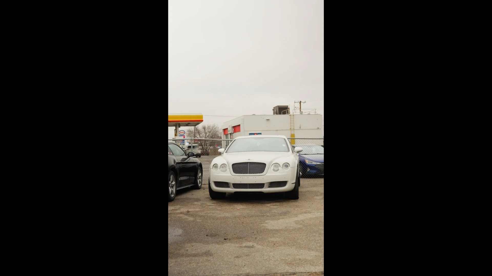 Front view of a white luxury sedan parked between a black car and a blue car in a lot, with a chain-link fence, buildings, and a gas station in the background under an overcast sky.