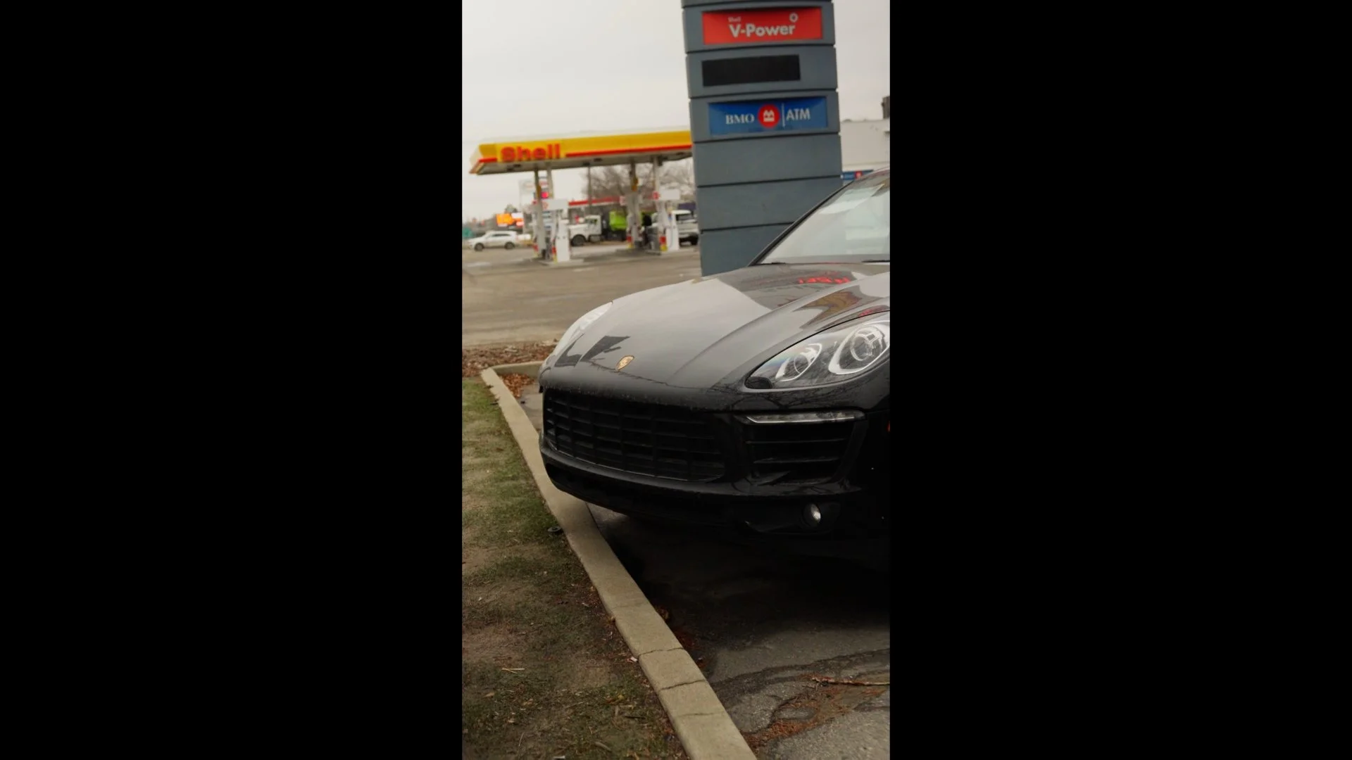 Black Porsche parked at a gas station with Shell and ATM signs visible in the background on an overcast day.