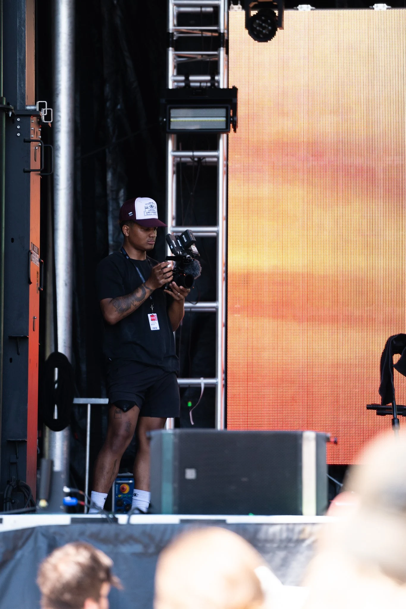 A person standing on a stage, holding a camera, with a large orange and yellow digital screen behind. The person is wearing a black shirt, black shorts, a baseball cap, and has tattoos and a lanyard with an ID badge.