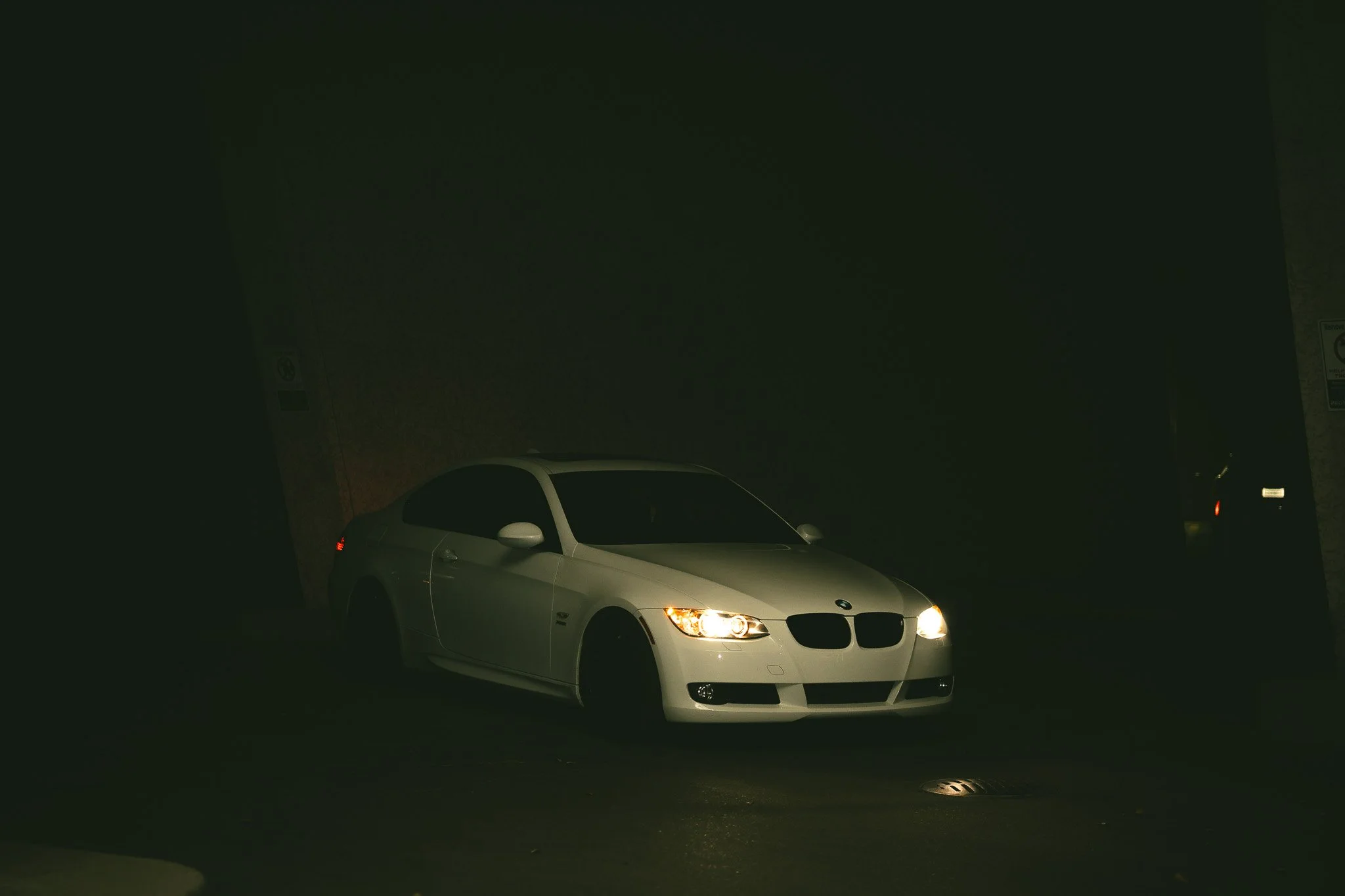 A white BMW car with its headlights on parked in a dark area.