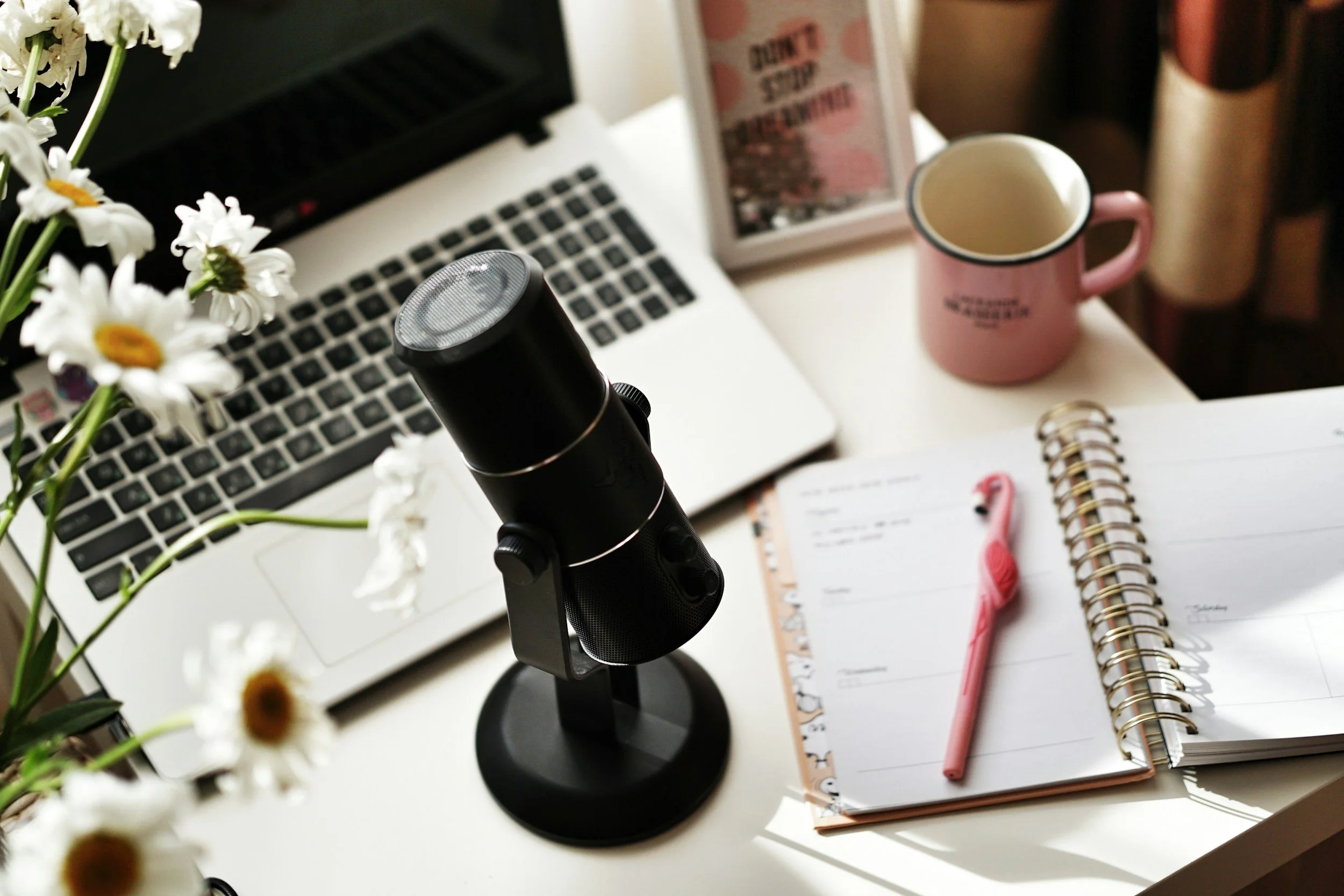 A workspace with a laptop, a microphone, a pink mug, a pink pen, a planner, and flowers on a white desk.