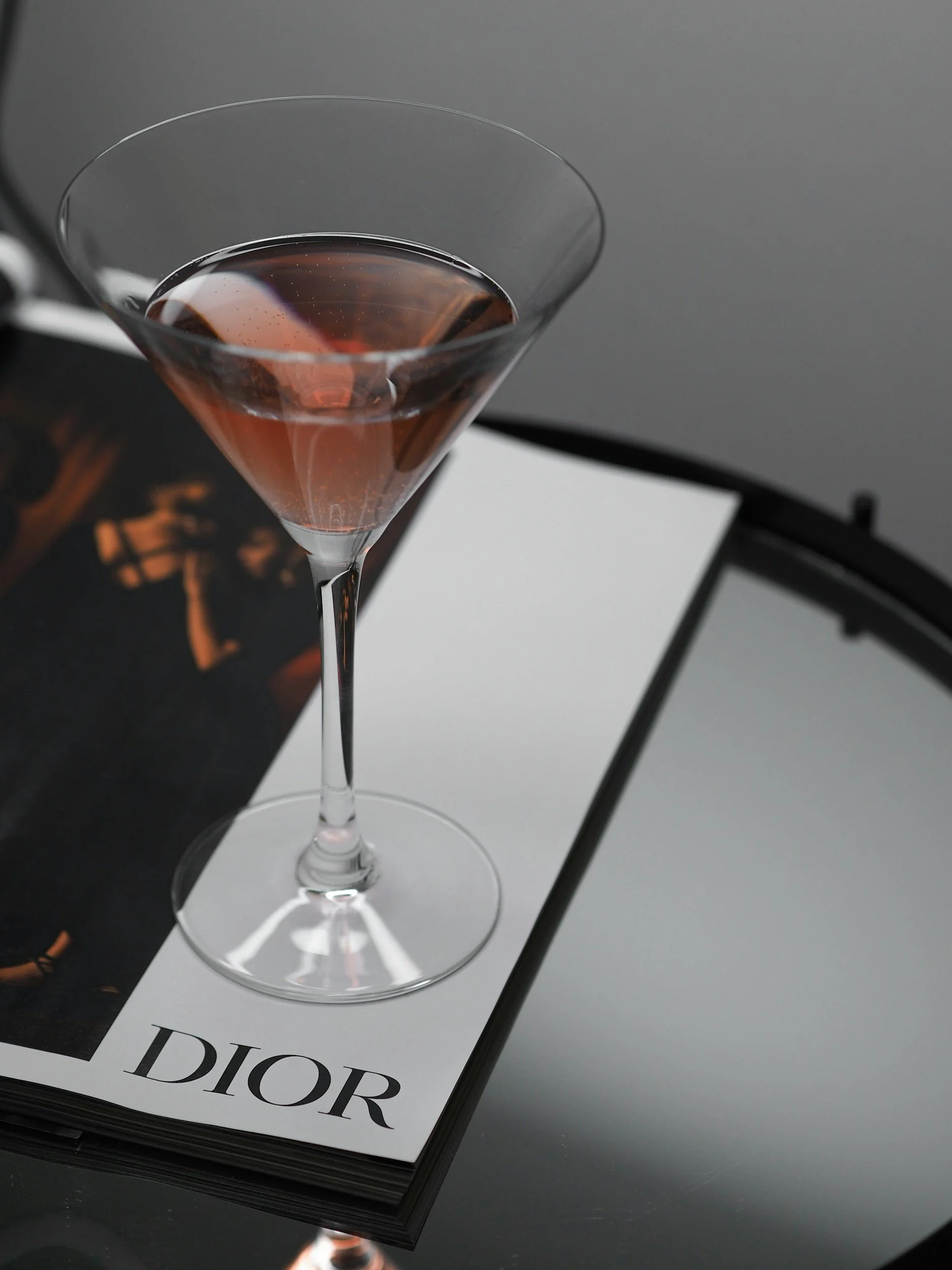 A martini glass with a pinkish-brown cocktail on top of a Dior magazine on a black glass table.