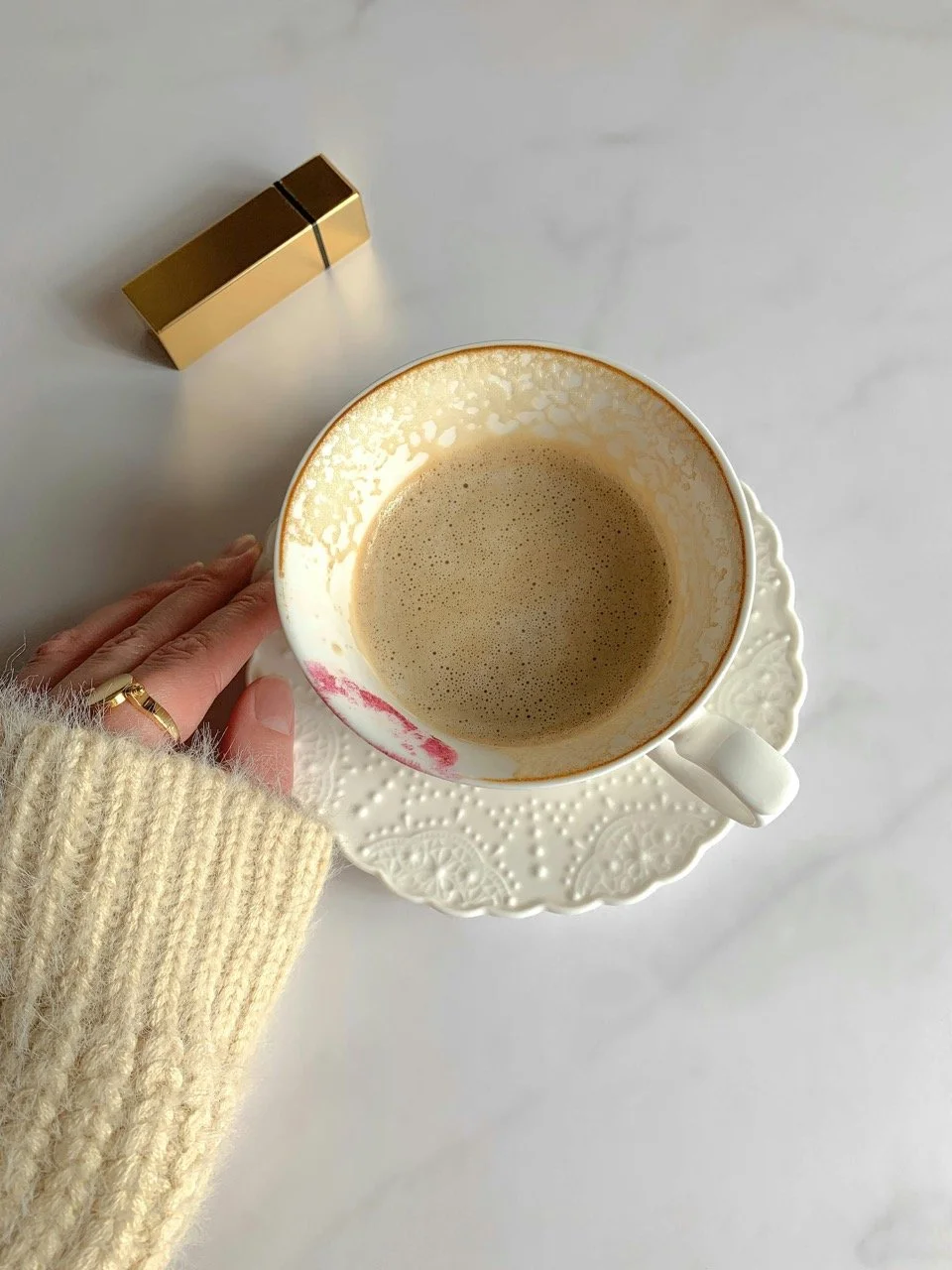 A cup of coffee on a white decorative plate, with a person's hand wearing a cream-colored sweater and a gold ring on their finger, resting nearby on a white surface; a gold rectangular object is in the background.