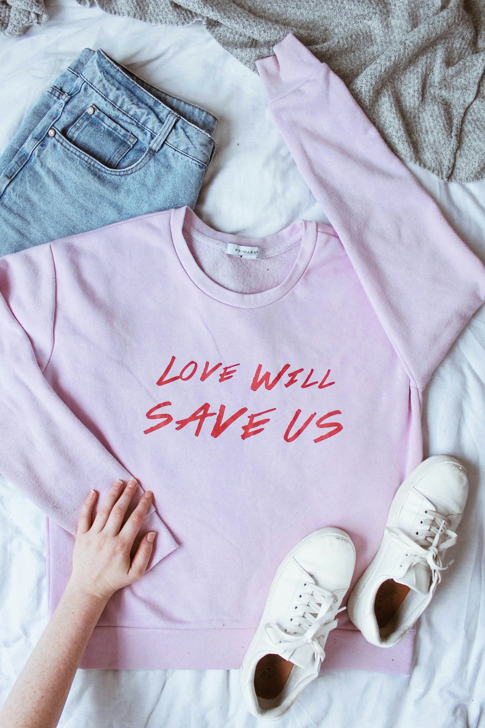 A pink sweatshirt with red text that says 'Love Will Save Us,' a pair of white sneakers, light blue jeans, and a grey sweater laid out on a white surface.