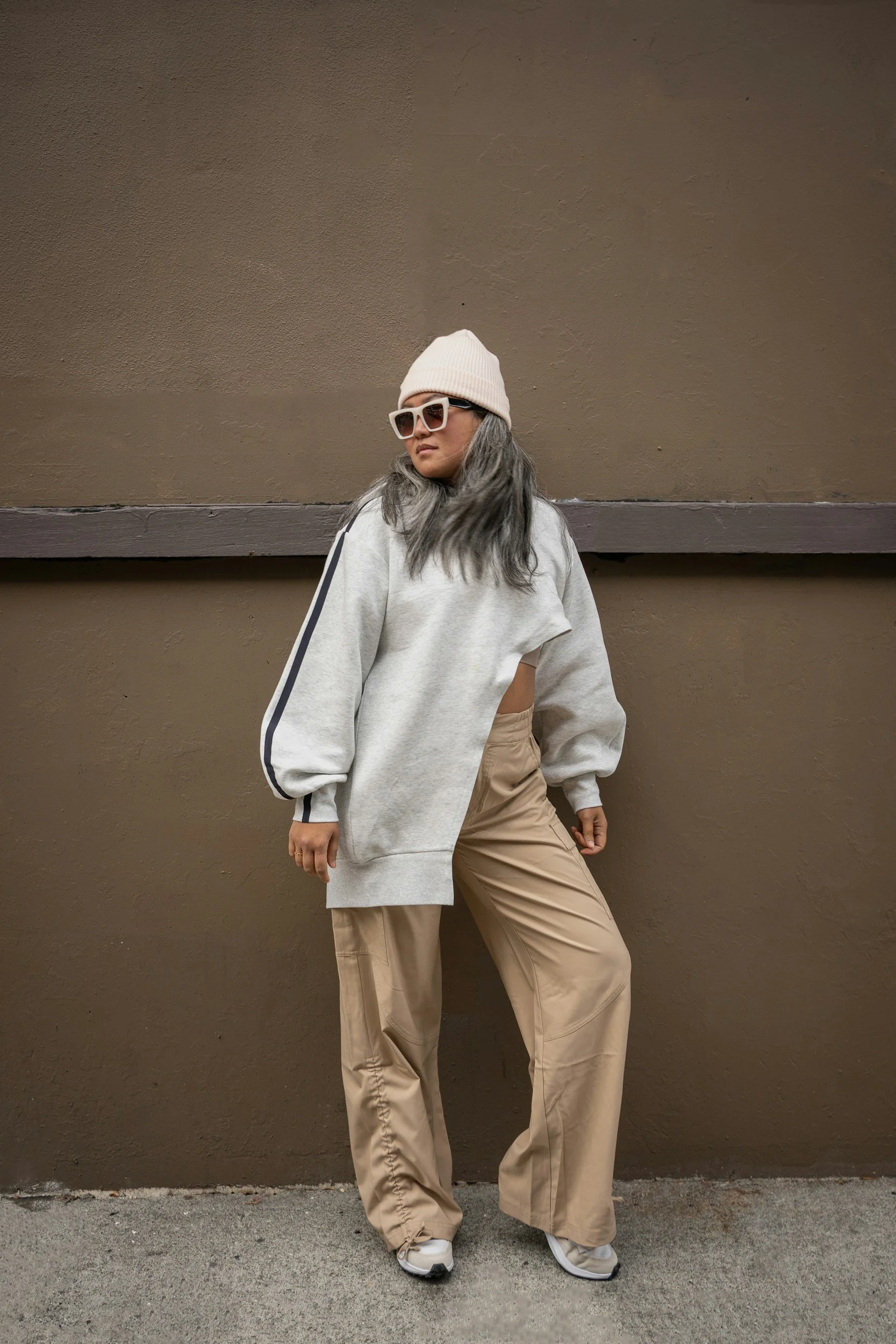 Woman with gray hair in beige wide-leg pants, gray sweatshirt, white beanie, and sunglasses posing against a brown wall.