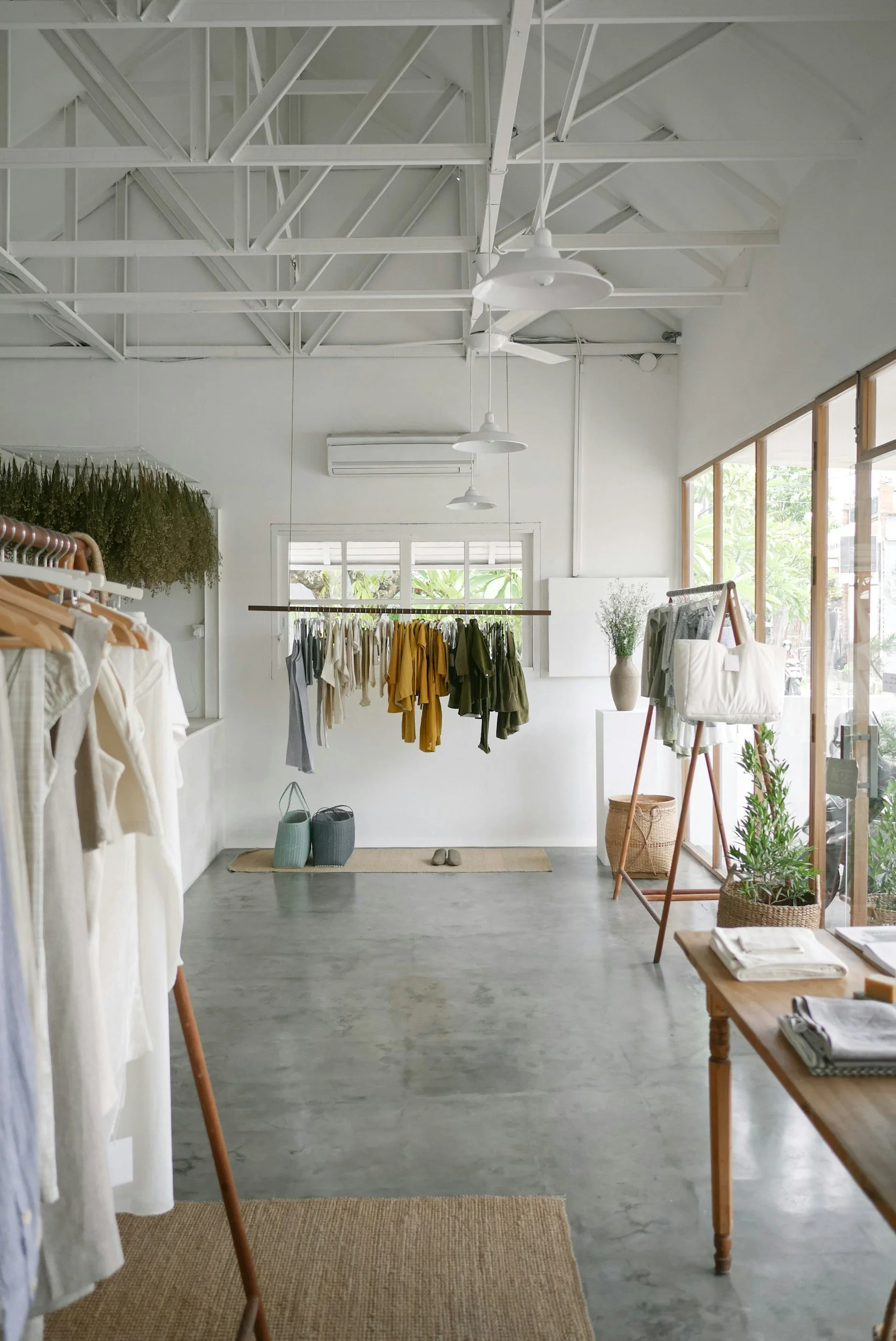 Interior of a minimalist clothing boutique with white walls, concrete floor, and large windows. Clothing items in neutral and earthy tones are displayed on racks and shelves, with decorative plants and baskets adding natural elements to the decor.