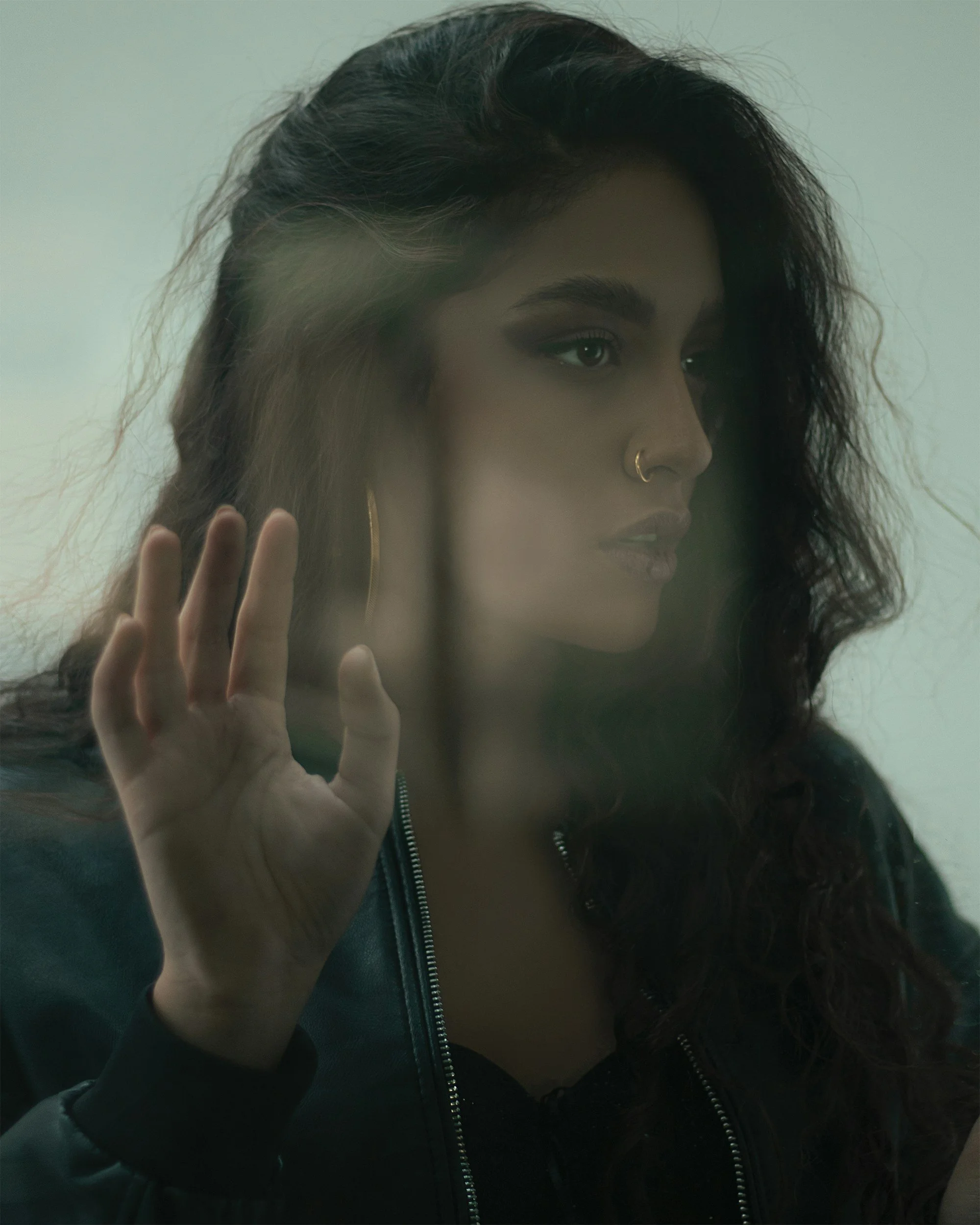 A woman with long, dark curly hair and hoop earrings looks at her reflection through a glass window, touching the glass with her hand.