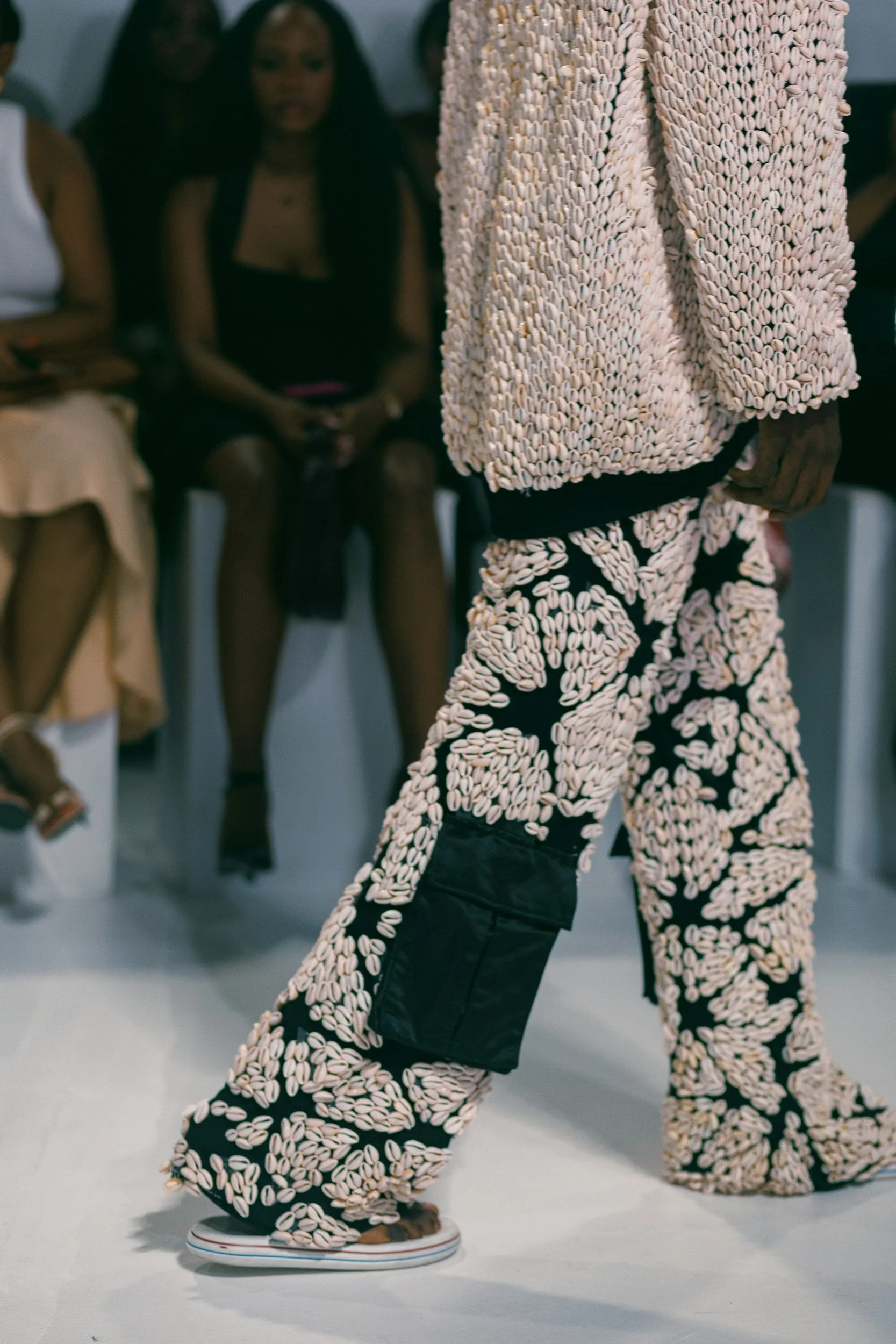 Person walking on runway wearing pants and a jacket decorated with numerous white seashells, with audience members seated in the background.