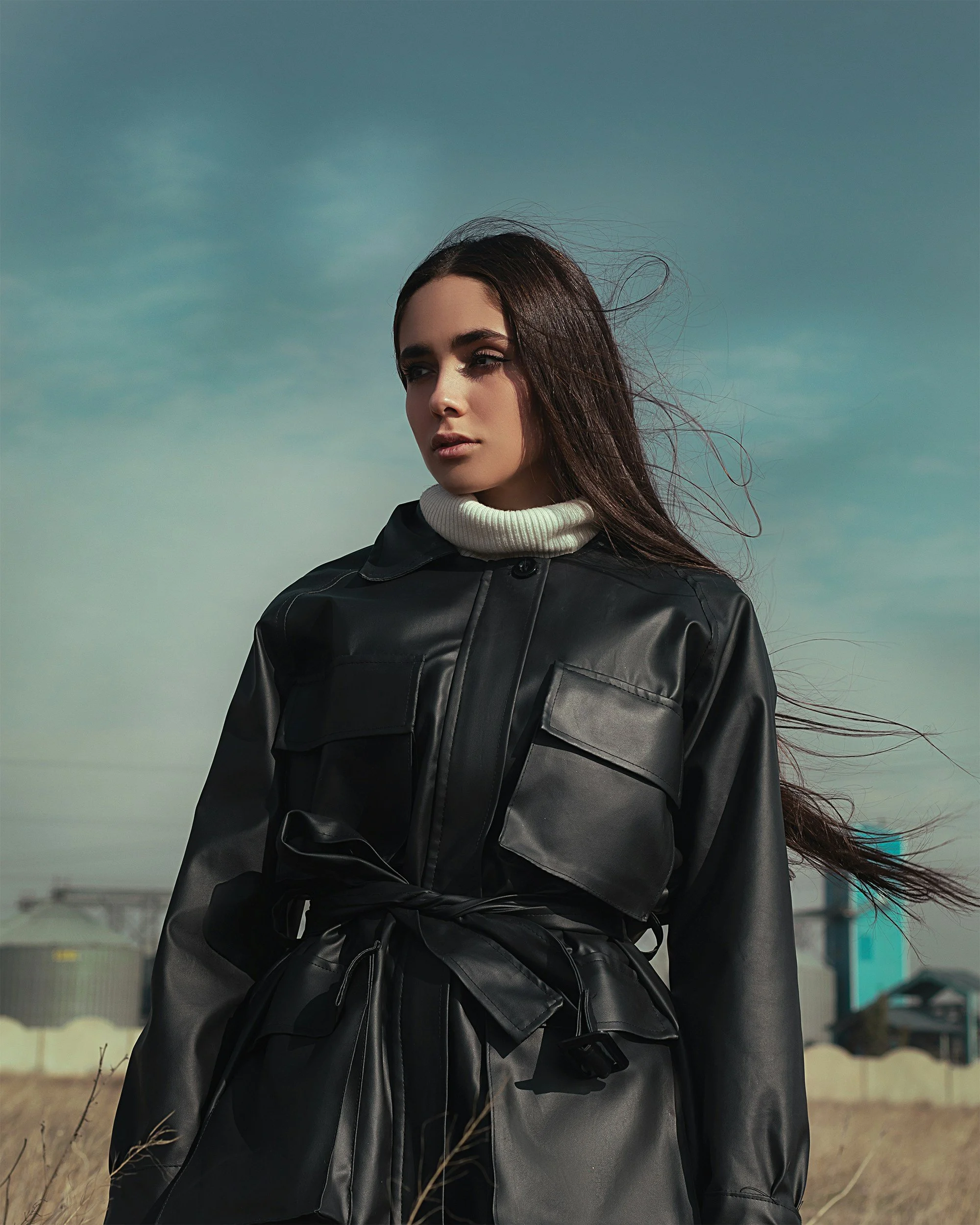 A woman with long dark hair standing outside on a cloudy day, wearing a black leather jacket with a waist belt and a white turtleneck sweater.