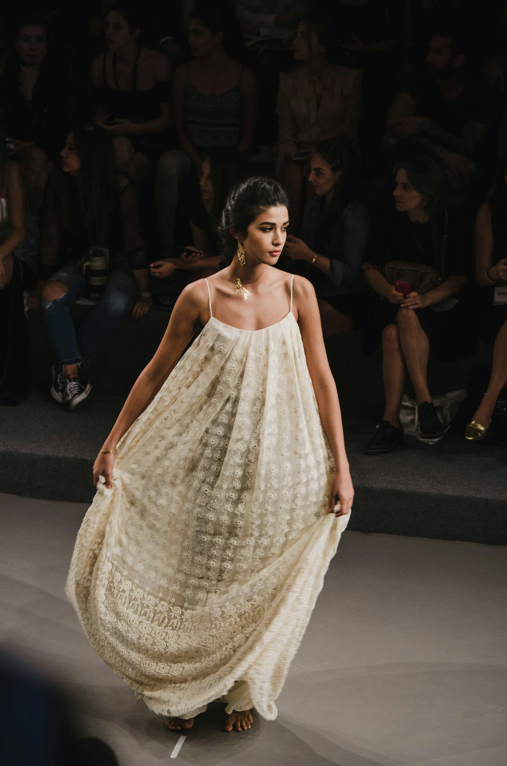 A female model walking on a runway wearing a flowy beige dress with spaghetti straps, accessorized with gold jewelry, with an audience watching in the background.