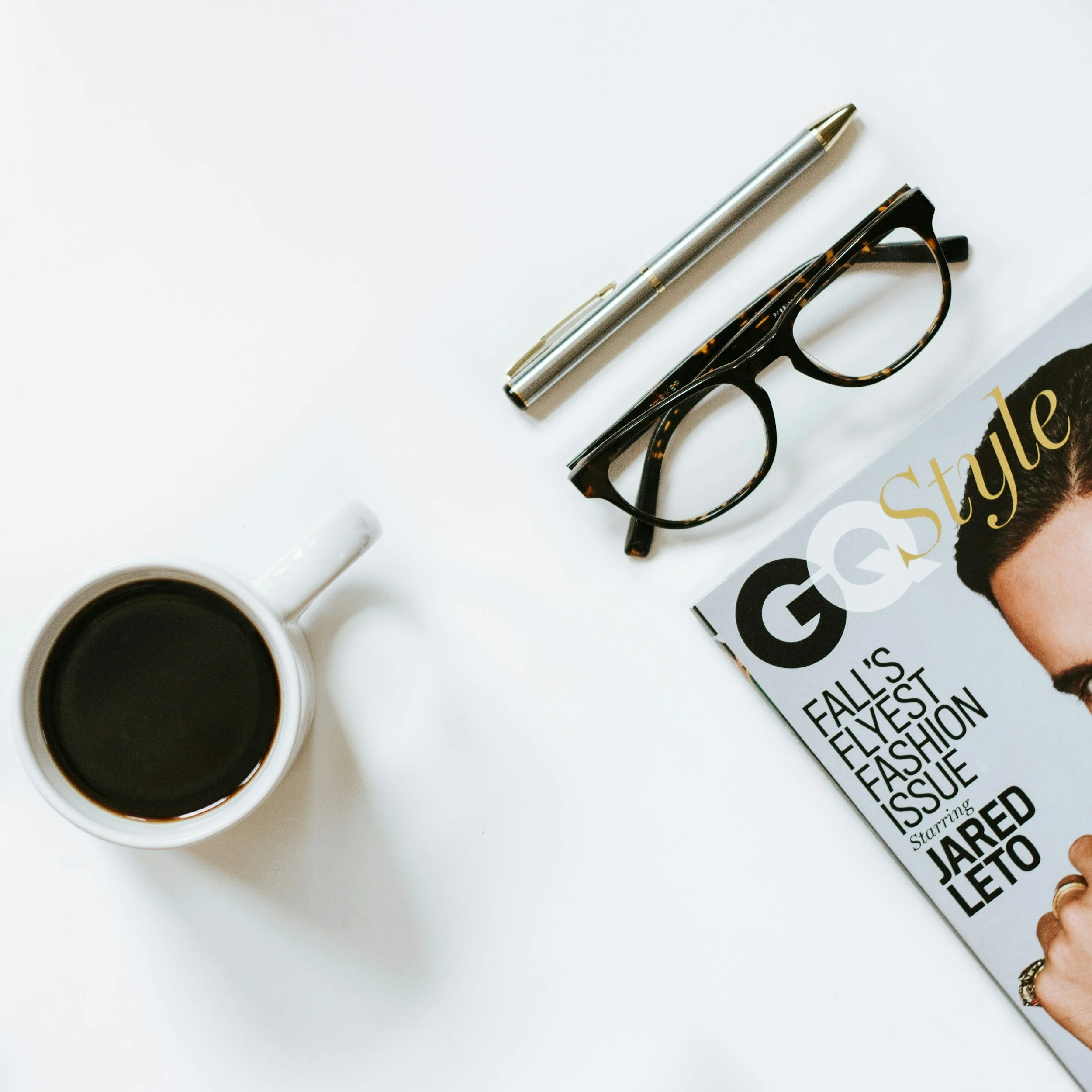 A white desk with a mug of coffee, a silver pen, tortoiseshell eyeglasses, and a GQ magazine featuring Jared Leto on the cover.