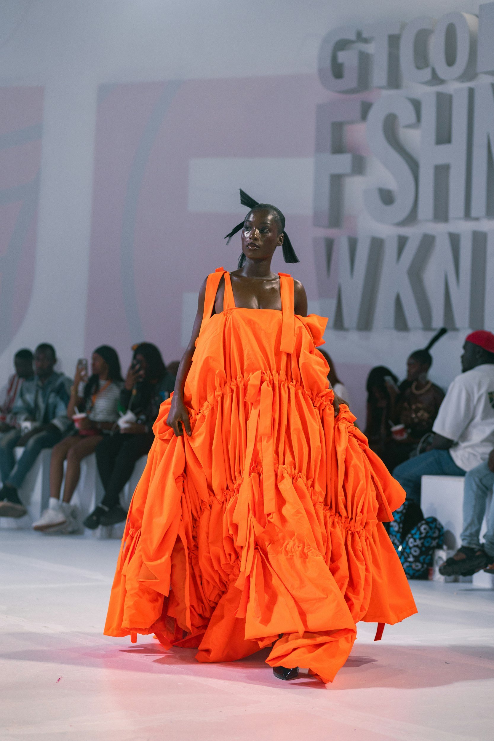 A model walking on a runway in a bright orange, voluminous dress with gathered and ruched details, during a fashion show. The background includes an audience and a large screen with stylized letters.