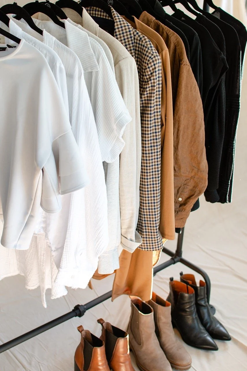 Clothing rack with white, beige, and black shirts and blouses and brown and checkered patterned garments. Shoes below include brown, gray, and black ankle boots.