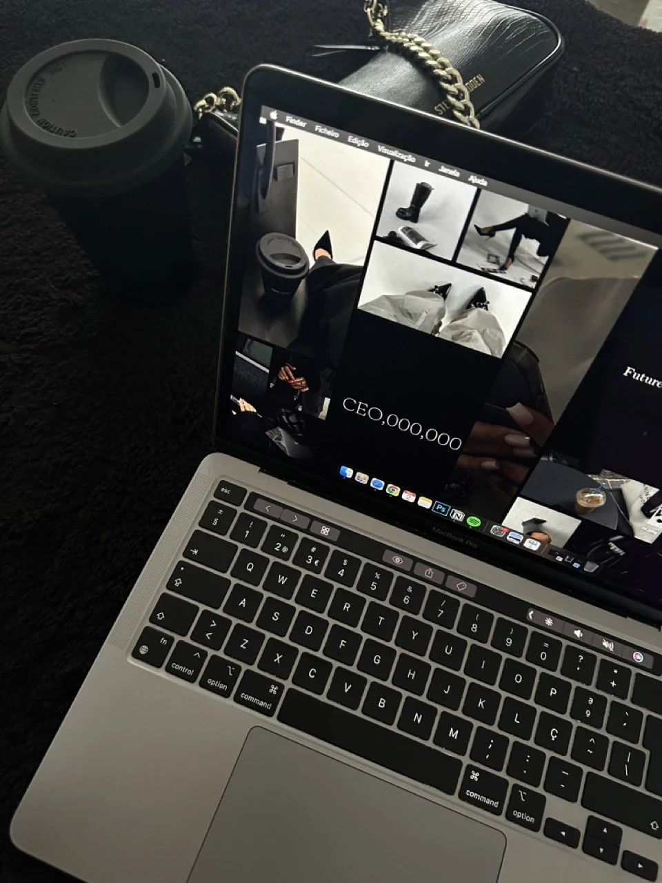 Close-up of a laptop with a website or presentation on the screen, a black travel mug, a black purse with a gold chain, and a disposable coffee cup on a dark textured surface.