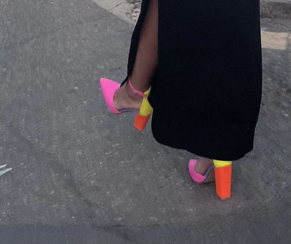 Close-up of a person's feet wearing neon pink and orange high-heeled shoes, with a black skirt and neon yellow accents on the shoes, walking on a paved surface.