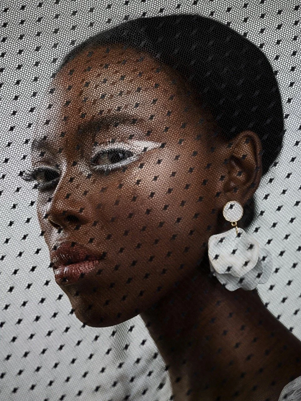 Close-up portrait of a woman with dark skin, wearing makeup with white eyeliner, and a large white earring, behind a dotted mesh fabric.