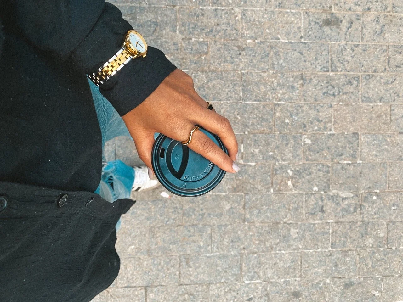 A person holding a blue disposable coffee cup on a sidewalk, wearing a black jacket, a gold watch, and a gold ring.