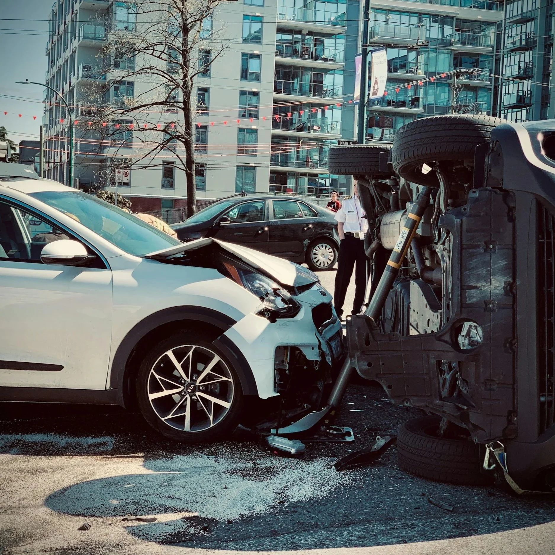 A car accident involving a white sedan and an overturned vehicle on an urban street with high-rise buildings in the background. Emergency personnel are present, and debris is scattered on the road.