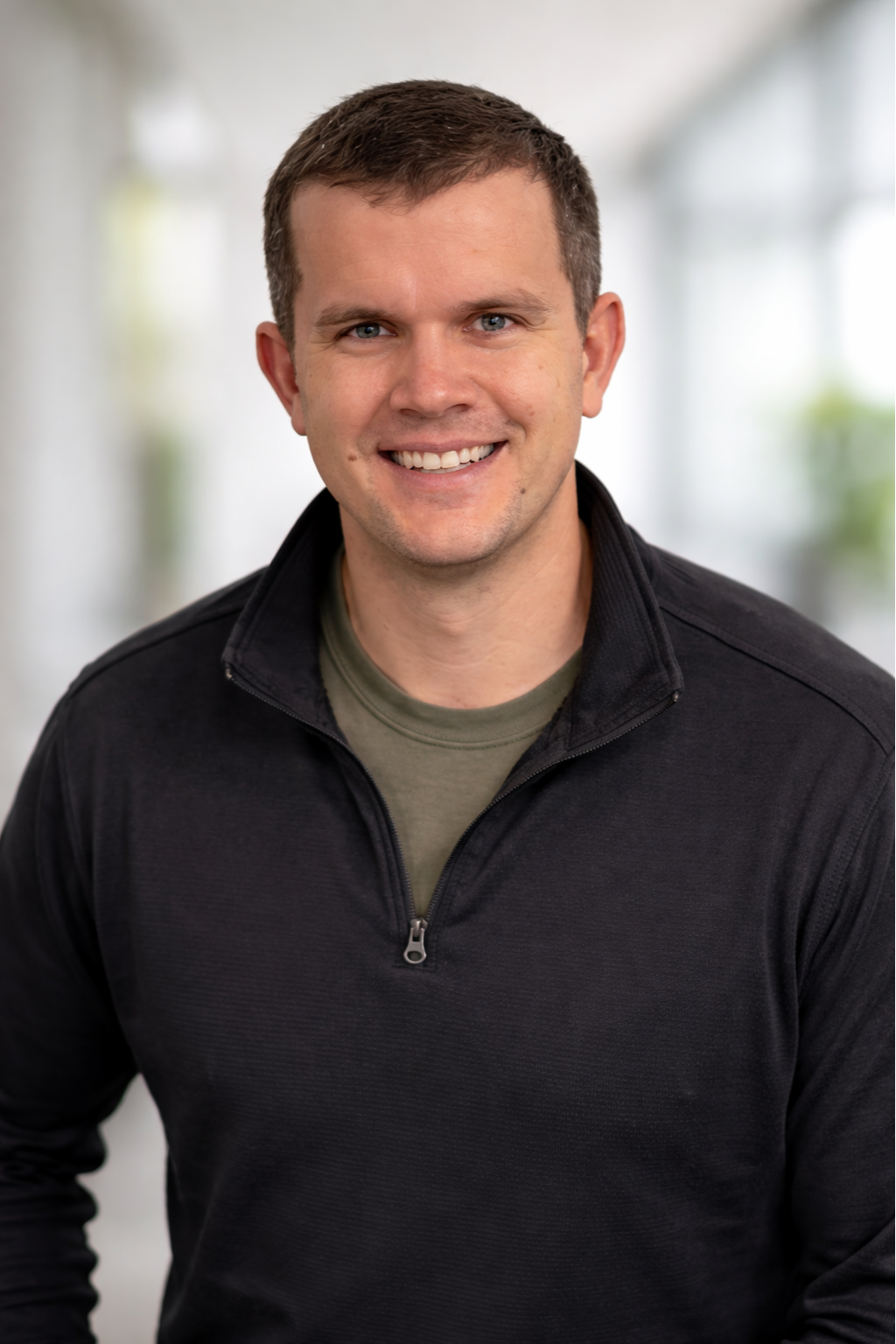 Headshot of smiling man with short brown hair, blue eyes, wearing a dark zip-up jacket and a green shirt, standing indoors with a blurred bright background.