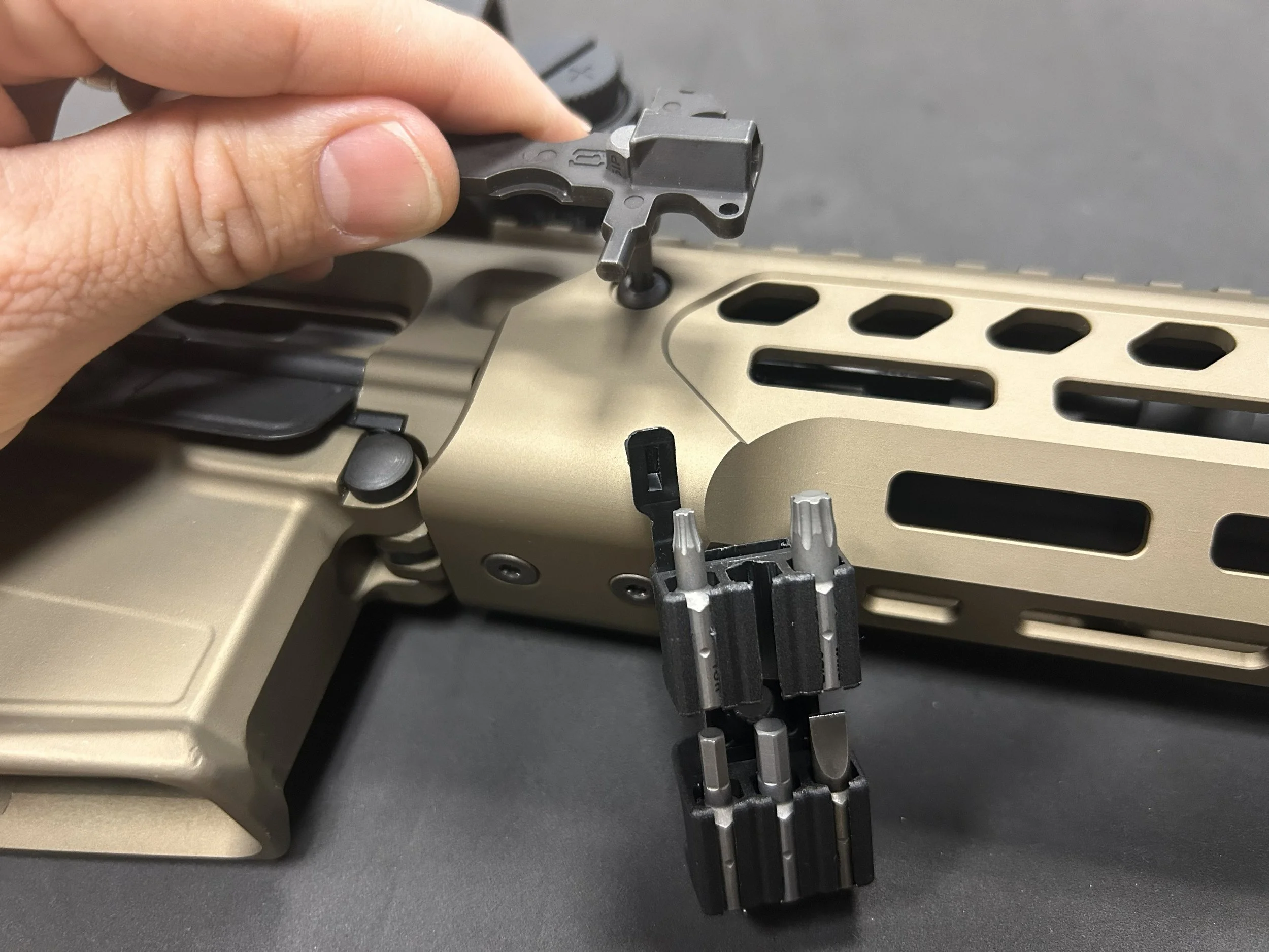 Close-up of a person installing or assembling a firearm part, with a tan-colored receiver, black components, and a person's hand holding a small metal piece.