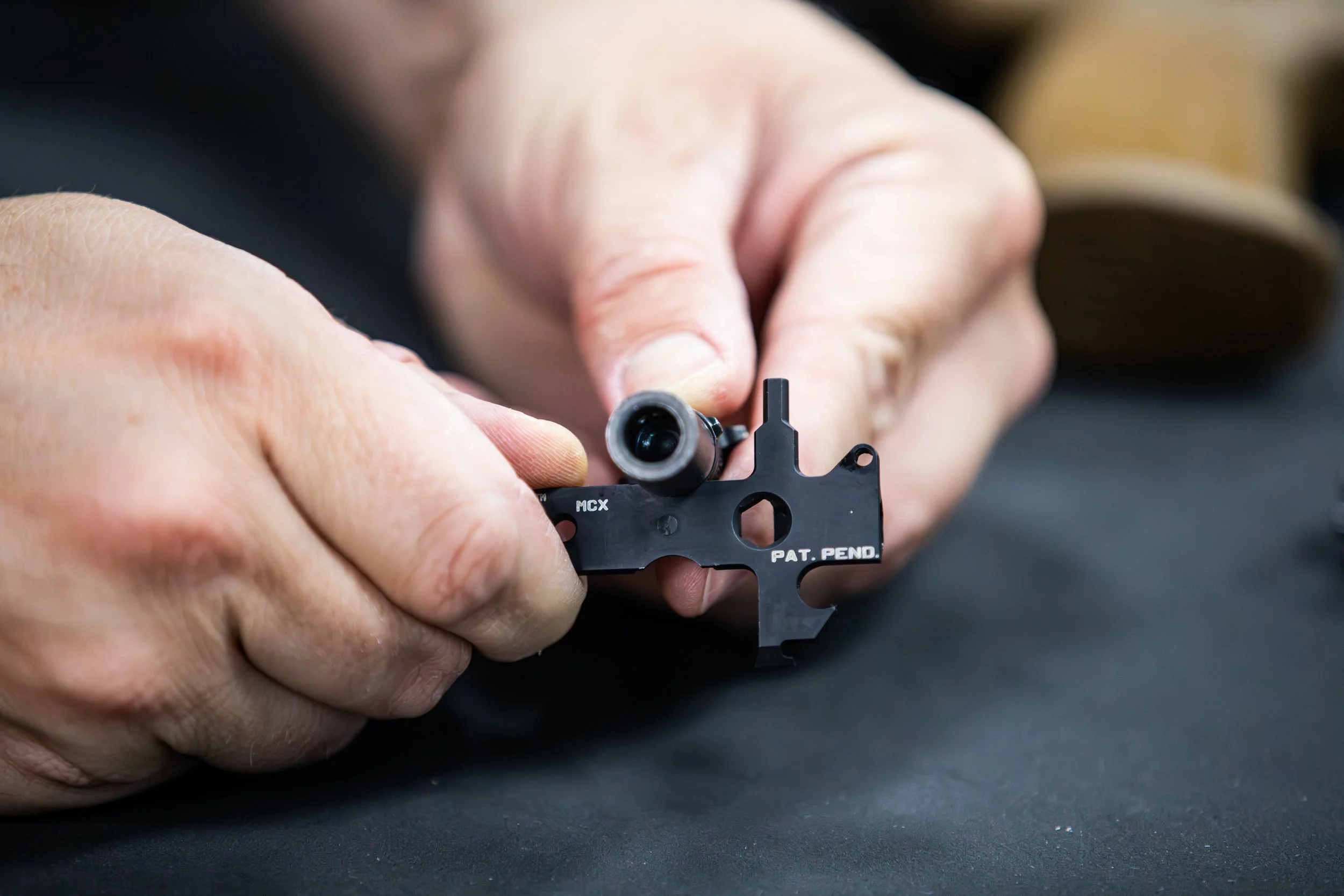 Hands holding a small black firearm part labeled 'PAT. PEND.' with a cylindrical barrel or attachment on it, placed on a dark surface.