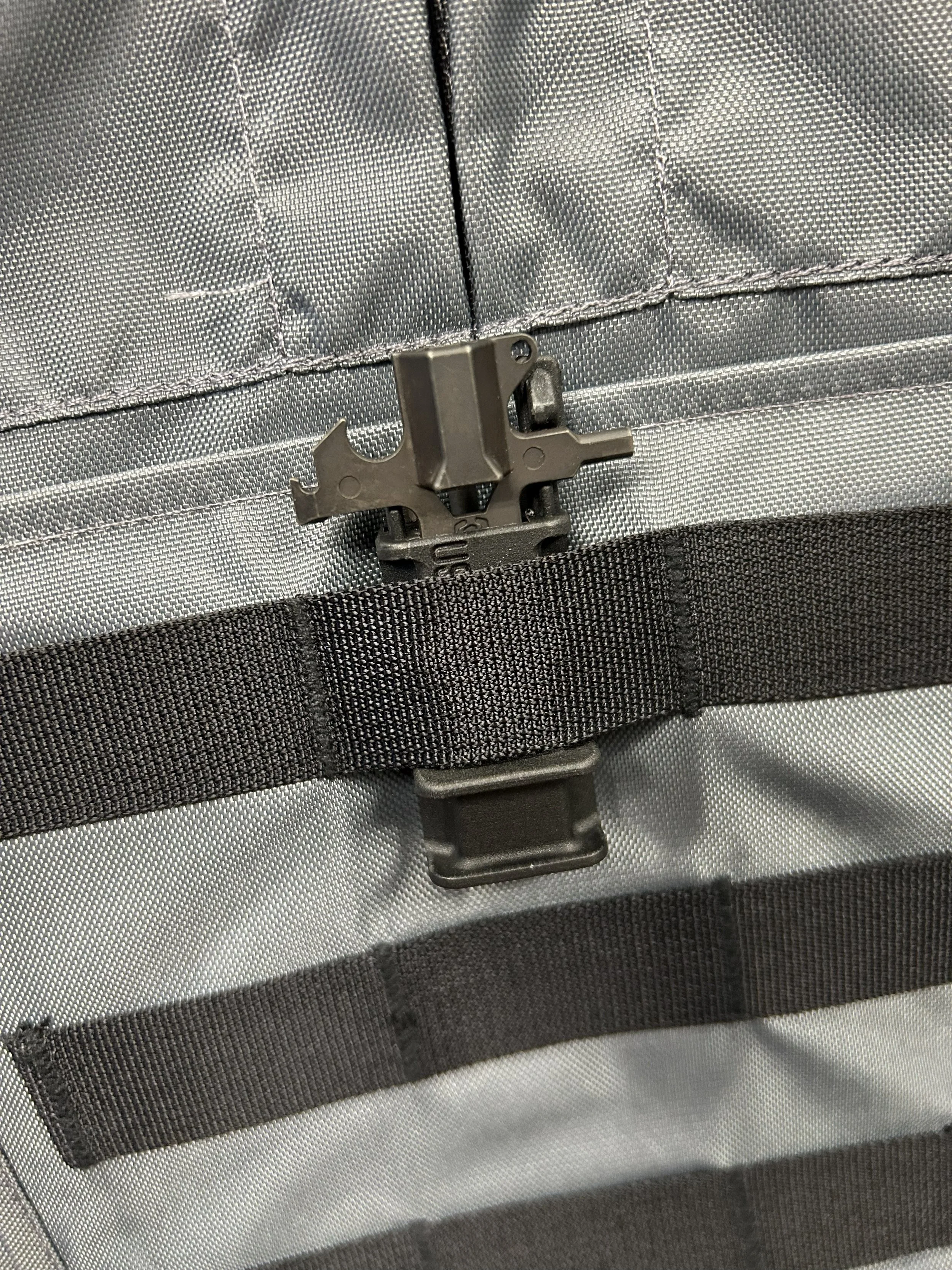 Close-up of a tactical backpack zipper with a metal lock tool attached to the zipper pull, on a gray and black backpack.