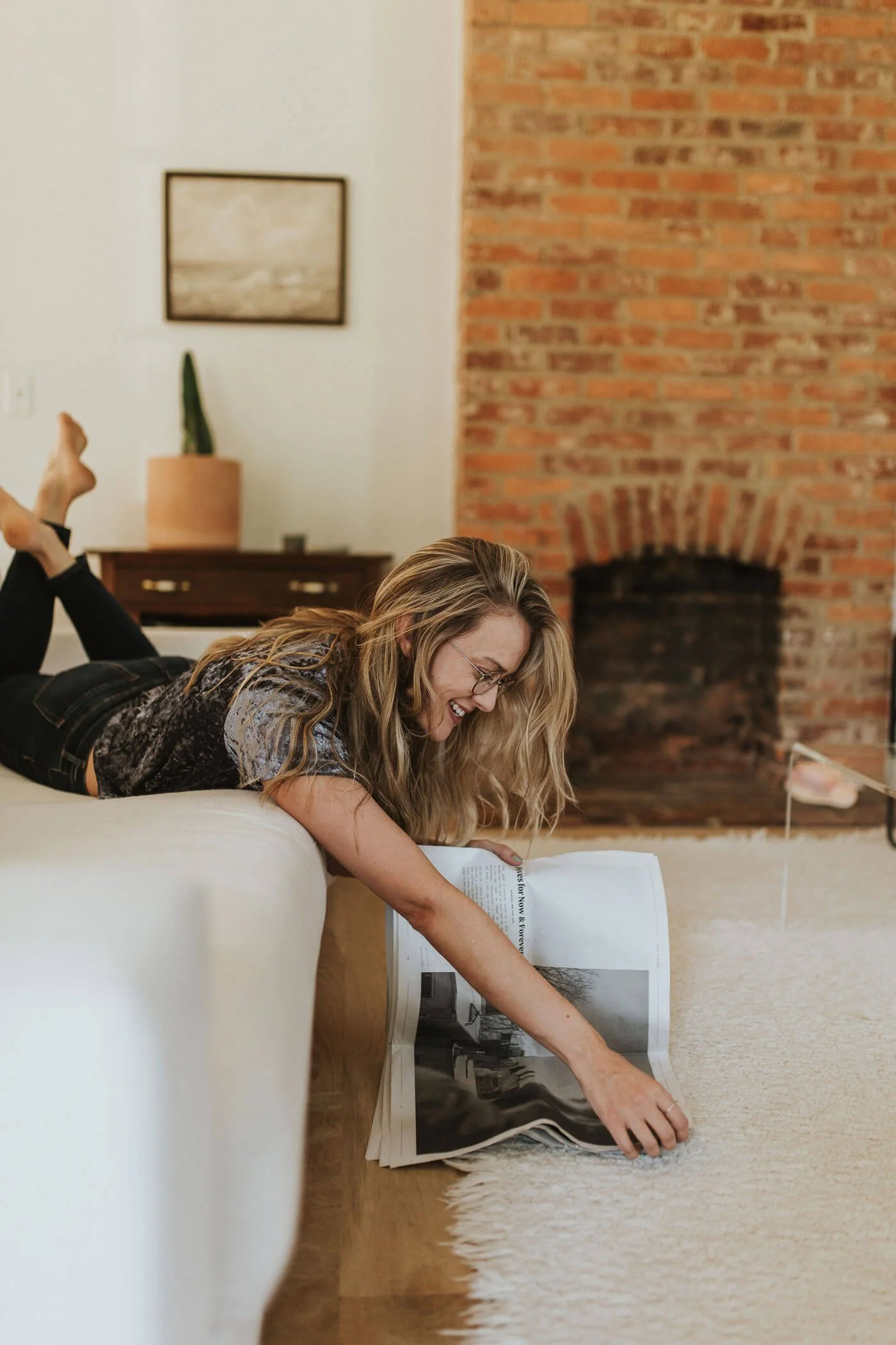 A woman with long blonde hair and glasses smiling while lying on her stomach on a white couch, reaching for a magazine on the beige carpeted floor in a living room with a brick fireplace and a small wooden table.