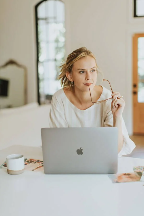 A woman with blonde hair sitting at a white desk with a silver MacBook, holding glasses, in a bright room with large windows.