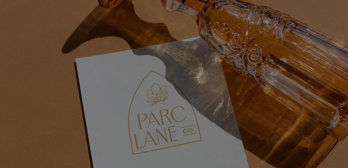 A white card with gold logo and text that reads "Parc Lane Co." is placed on a brown surface. Next to it, there is a clear glass decanter with intricate patterns, partially shown, casting reflections and shadows.