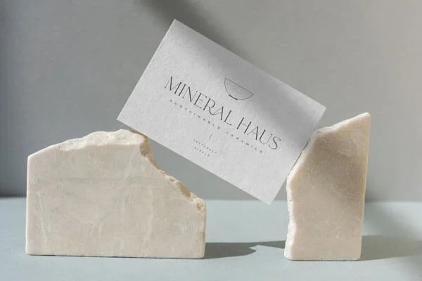 A white bar of soap with rounded edges and a smaller soap piece next to it, with a business card that reads 'Mineral Haus' leaning against the smaller soap piece.
