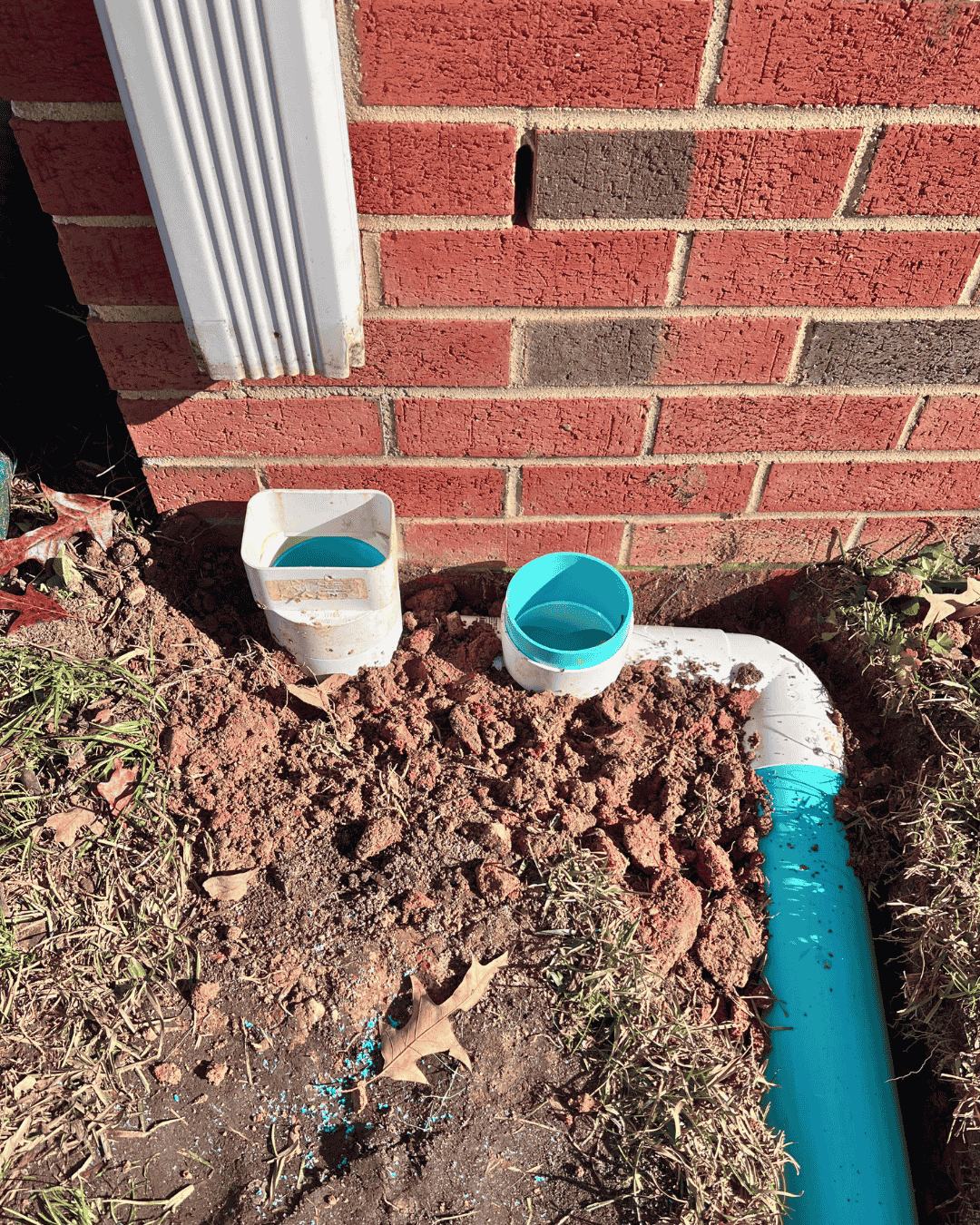 Downspout tie-in with solid PVC drainage line beside a brick foundation—roof runoff drainage work to move water away from the home in Cary, NC.