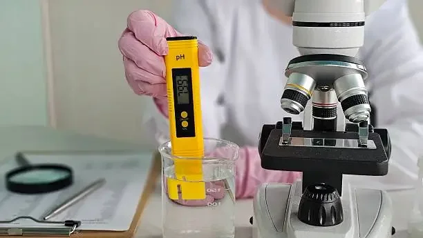 Scientist measuring pH levels of a liquid using a digital pH meter beside a laboratory microscope.