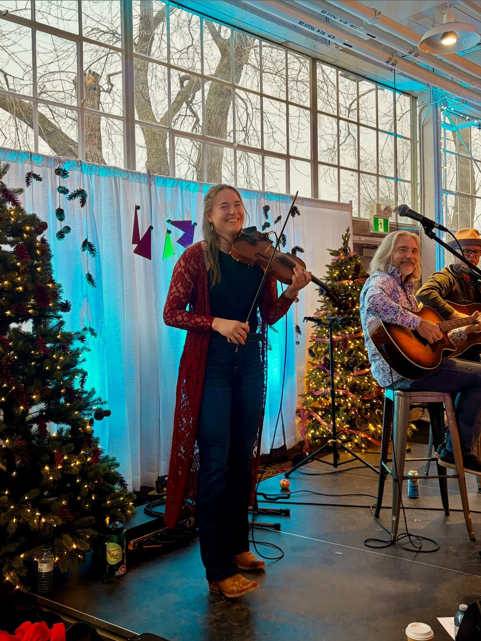 Musicians performing on stage during a holiday event with Christmas trees and holiday decorations, including wrapped lights and ornaments.