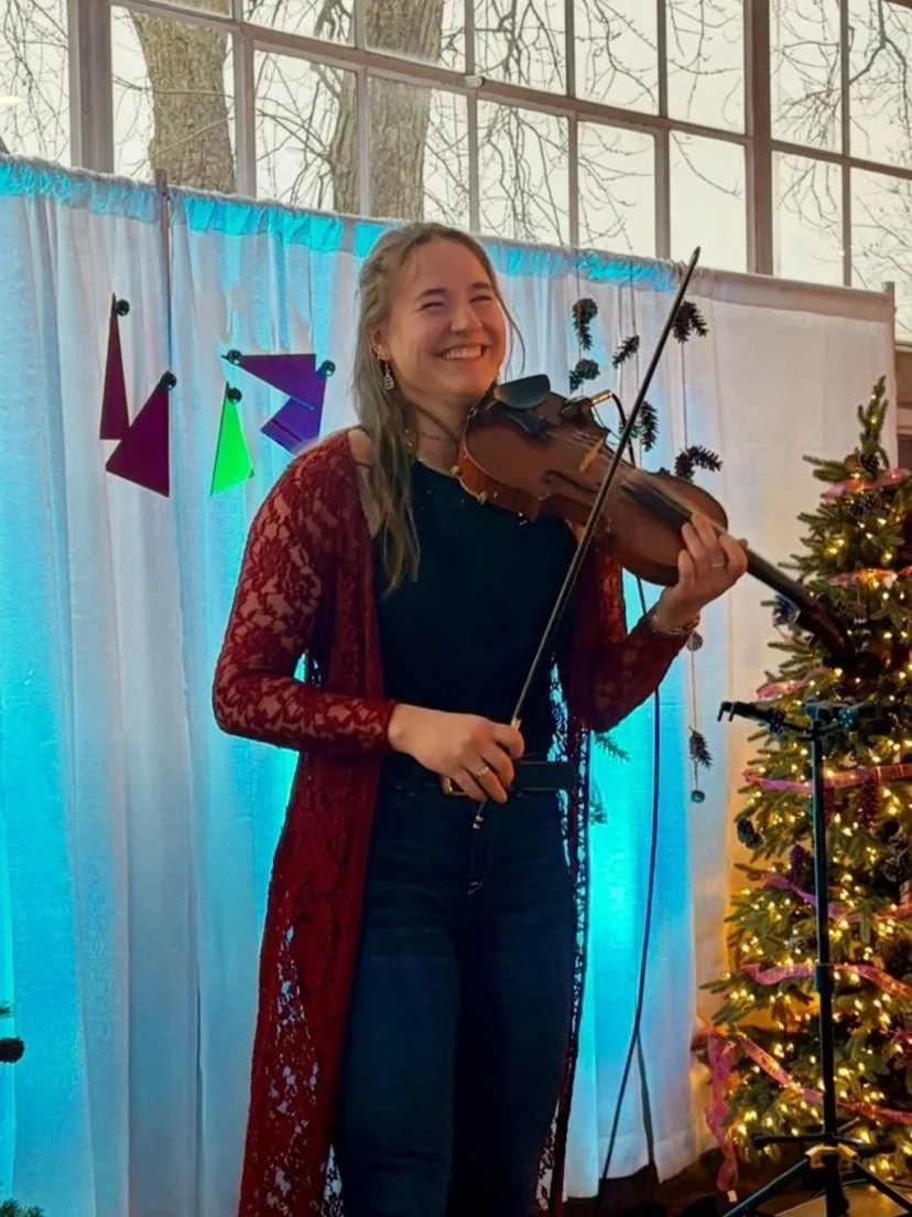 A young woman smiling and playing a violin at a festive indoor event next to a decorated Christmas tree. She is wearing a black top, dark jeans, and a red lace cardigan, with holiday-themed decorations in the background.