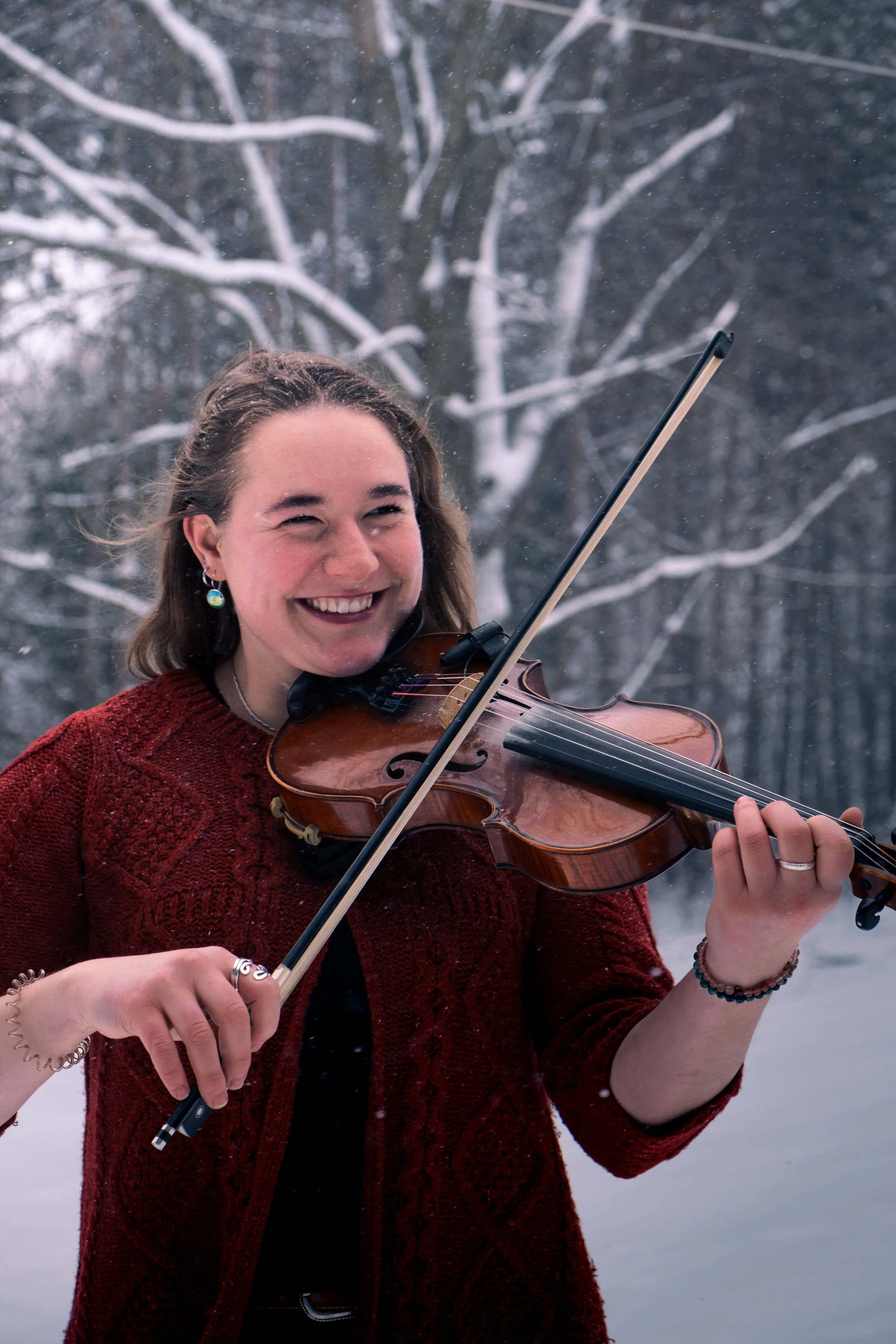 A young woman with long brown hair playing a violin outdoors in a snowy forest, wearing a red sweater and smiling.