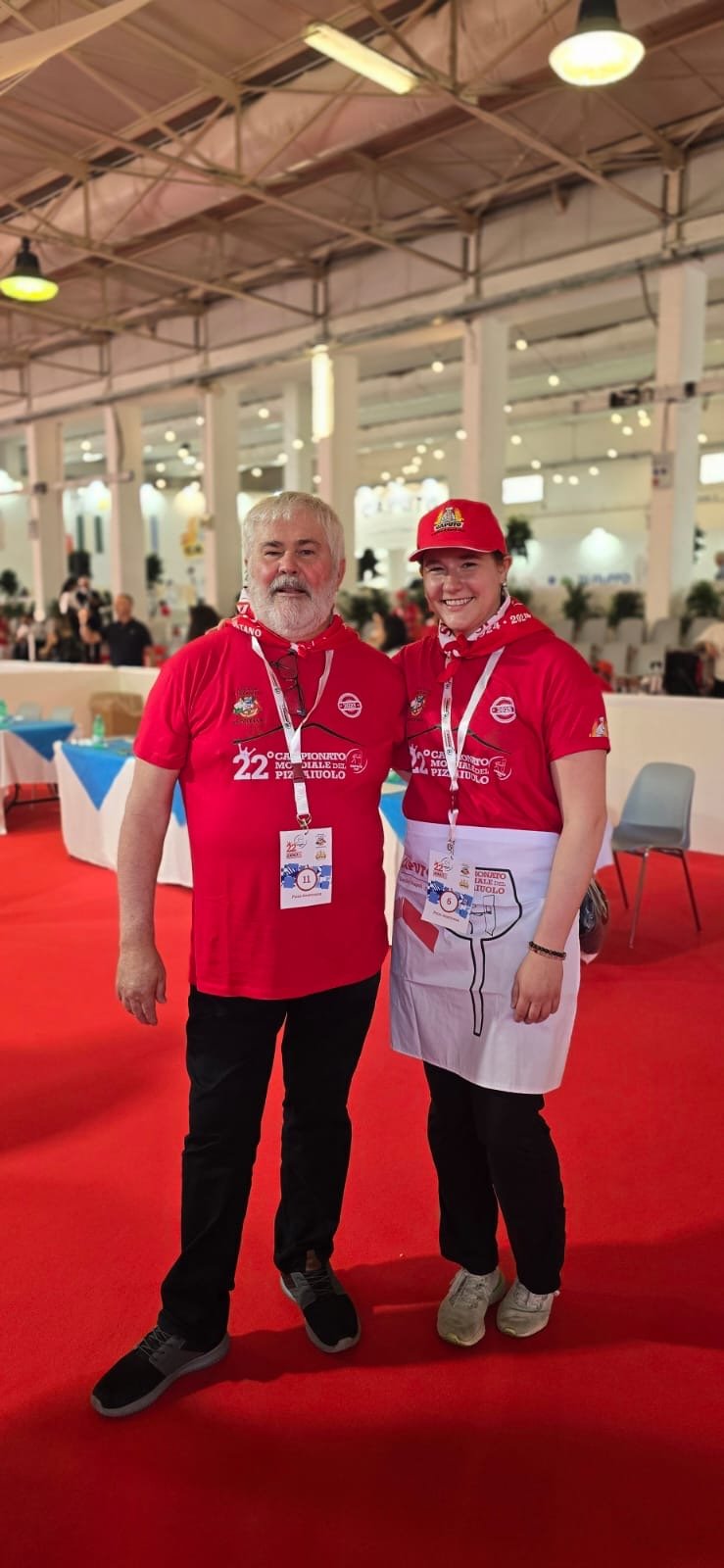 Two people wearing red shirts with event logos, standing inside a large indoor venue with red flooring and tables, participating in a culinary or sports event.