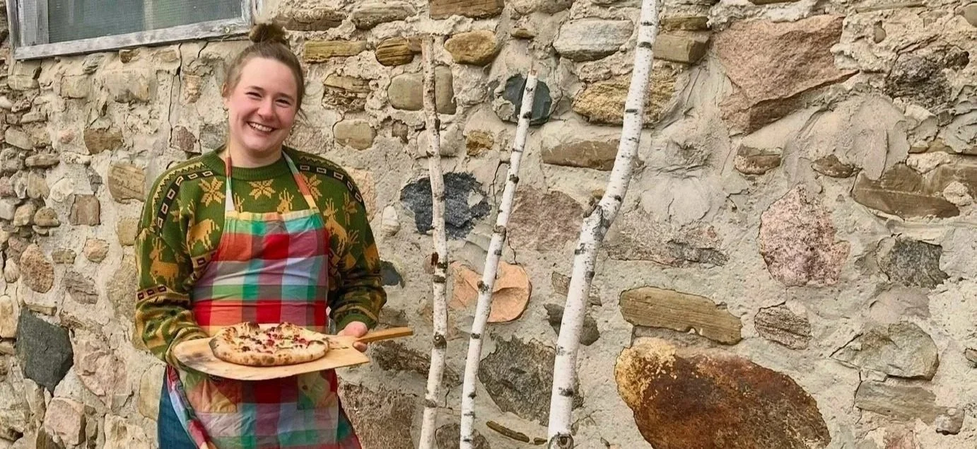 A woman holding a pizza on a wooden board, standing in front of a stone wall with birch trees.